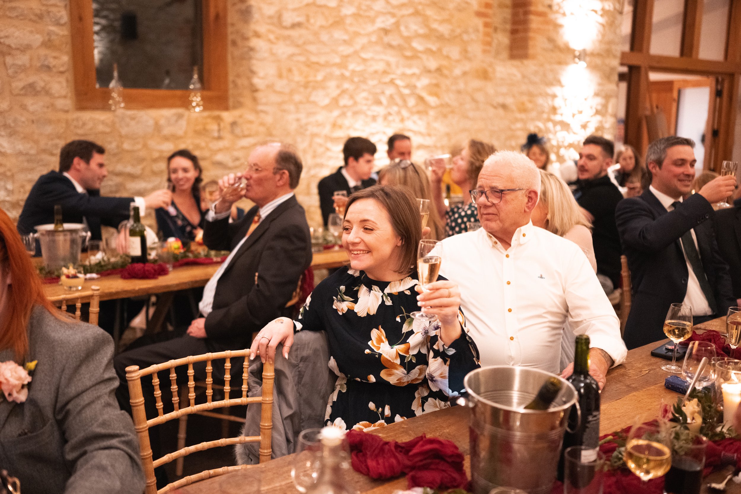 People at a celebration or dinner party holding glasses of champagne, sitting at long tables decorated with flowers, candles, and wine bottles in a rustic, warmly lit venue with stone walls.
