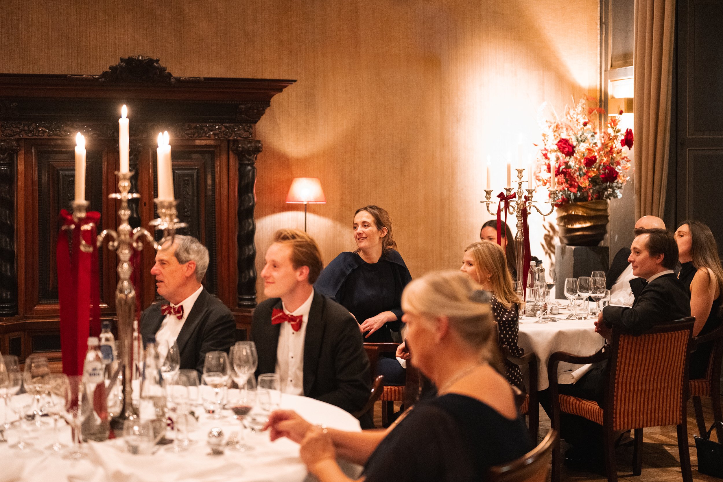 Guests seated at a formal dinner table in an elegant room with warm lighting, ornate dark wood furniture, candle centerpieces, and a woman standing and smiling.