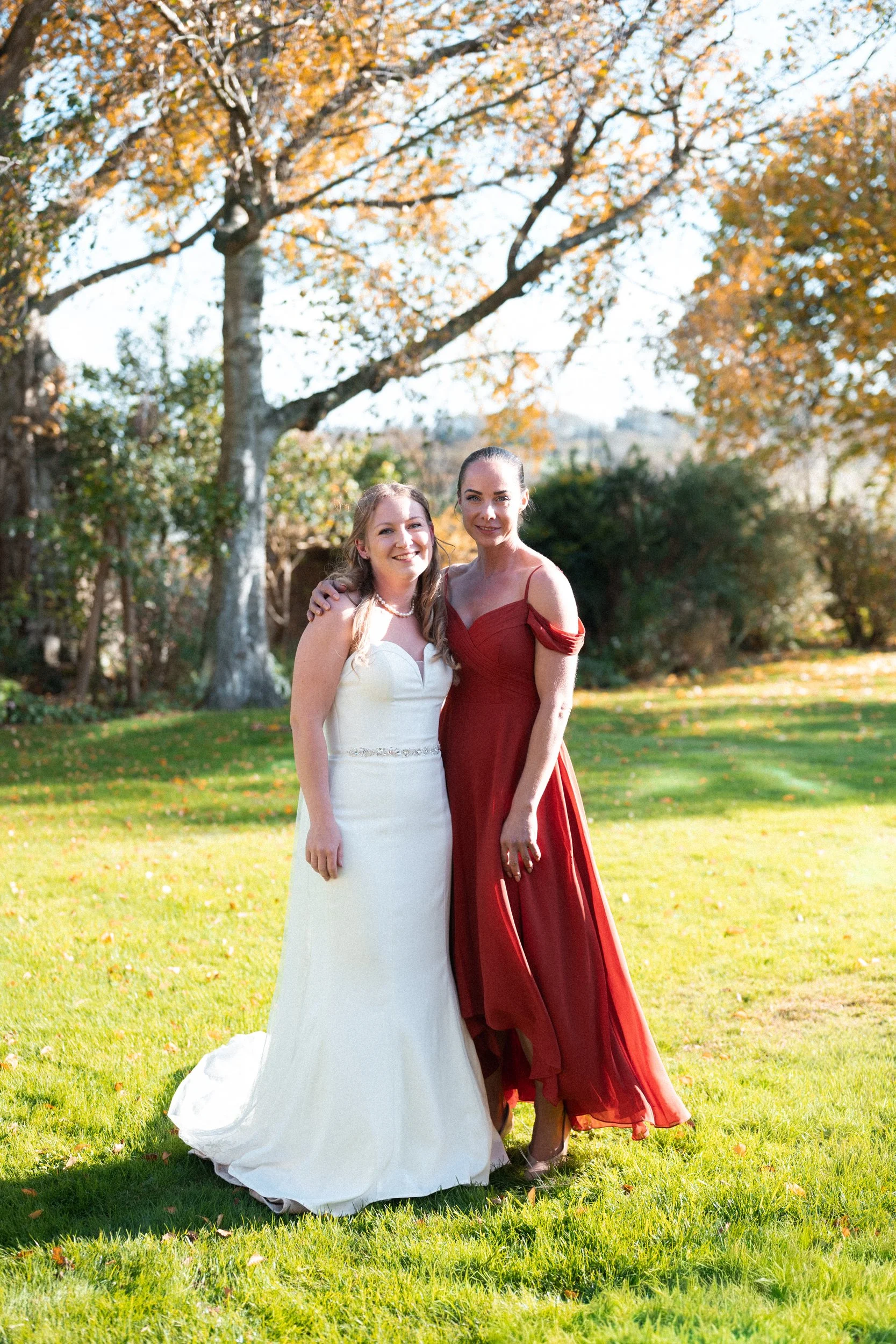 Two women standing outdoors on a sunny day, one in a white wedding dress and the other in a red evening gown, smiling at the camera surrounded by autumn trees and green grass.