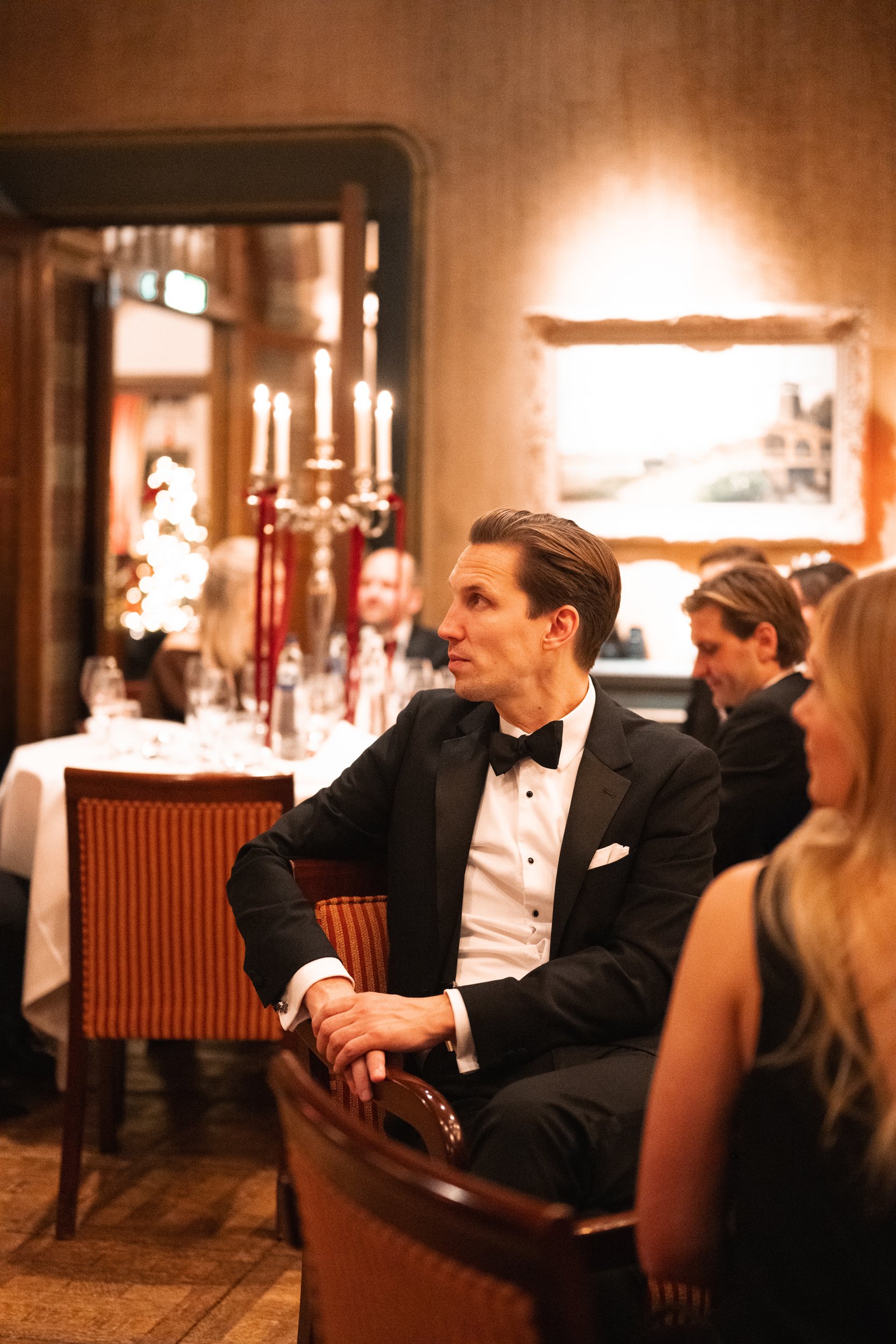 A man in a black tuxedo with a bowtie sitting in a decorated dinner setting, surrounded by other formally dressed guests, with a candelabra and candlelit table in the background.