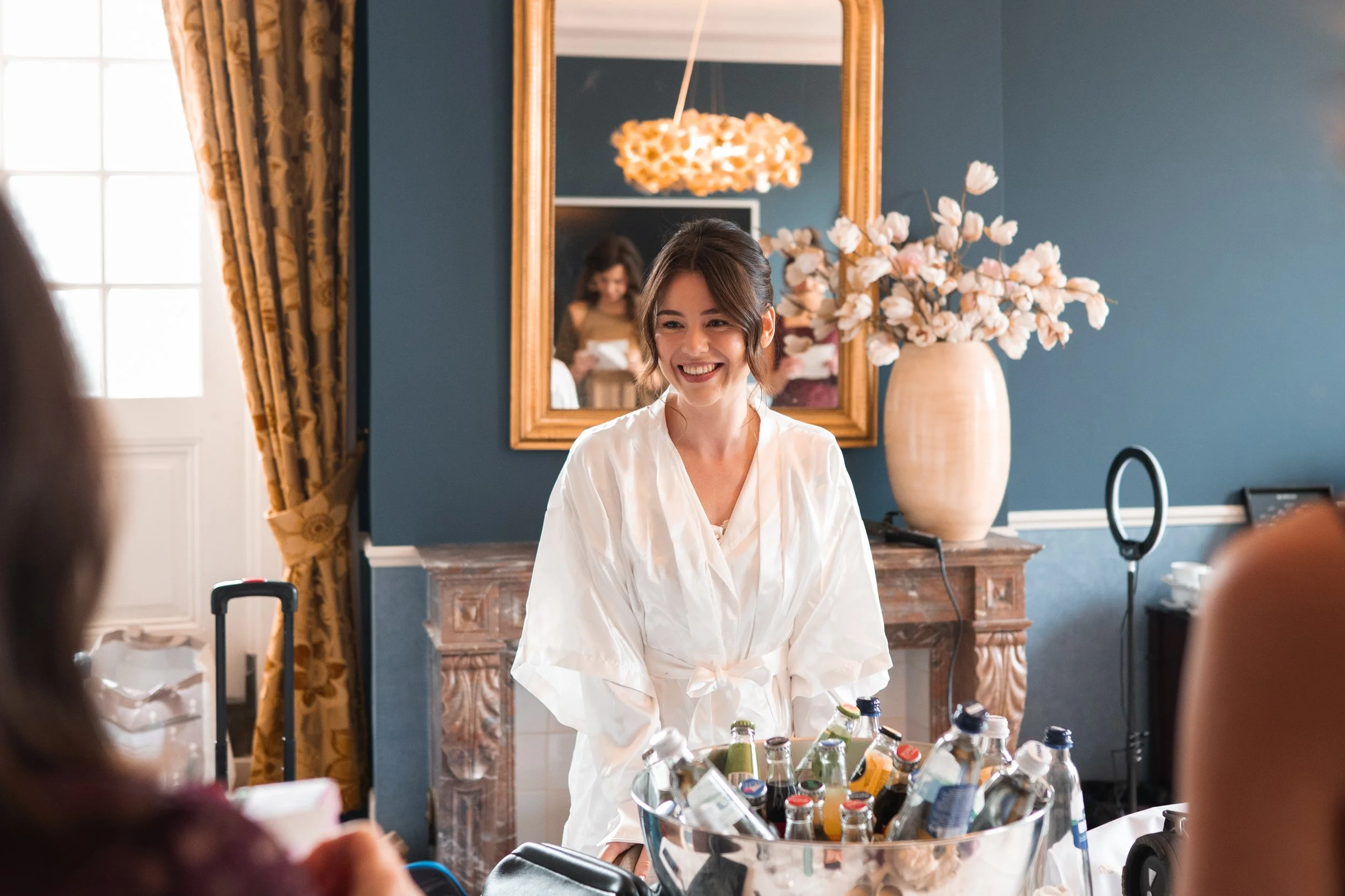 Woman in white robe smiling during a social gathering with bottles of beverages on the table, in a room with blue walls, a large mirror, a vase with white flowers, and gold curtains.