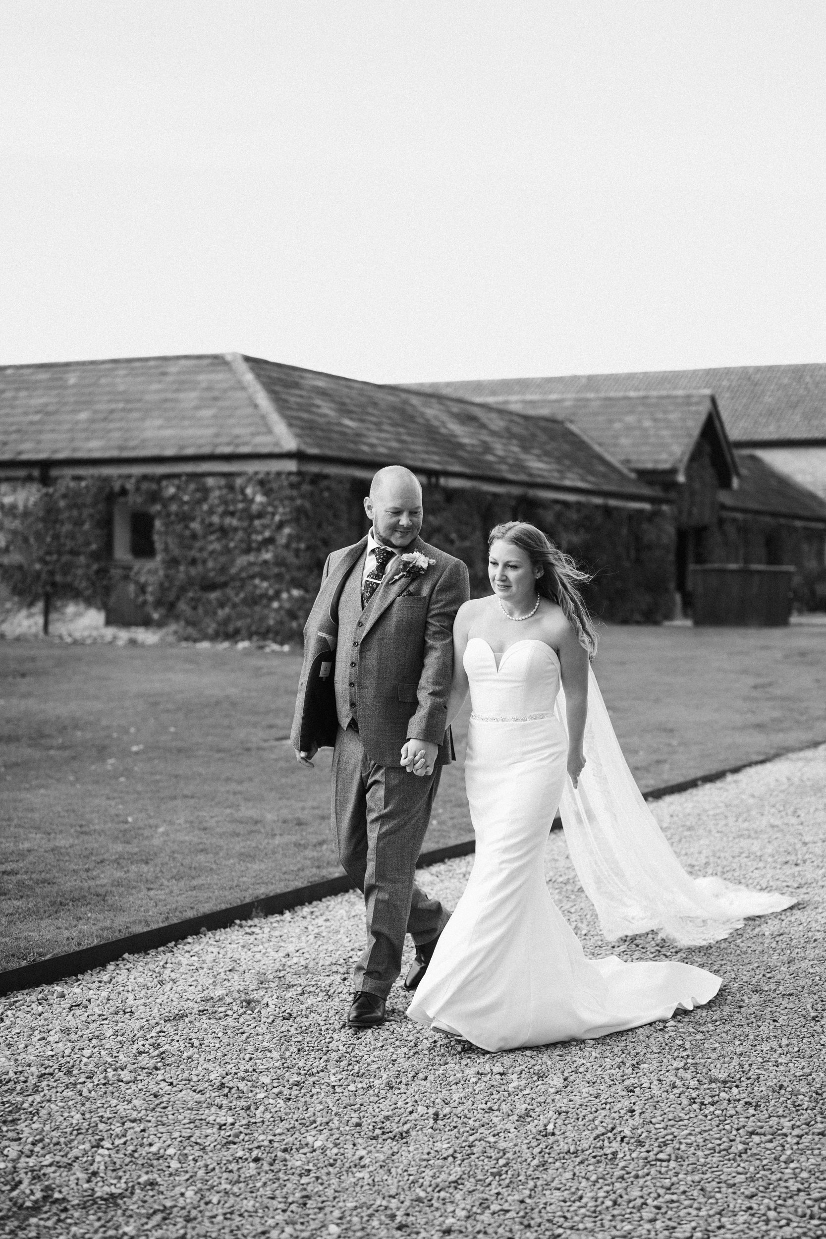 A black-and-white photo of a bride and groom walking hand-in-hand outdoors, with rustic buildings in the background, on a gravel path.
