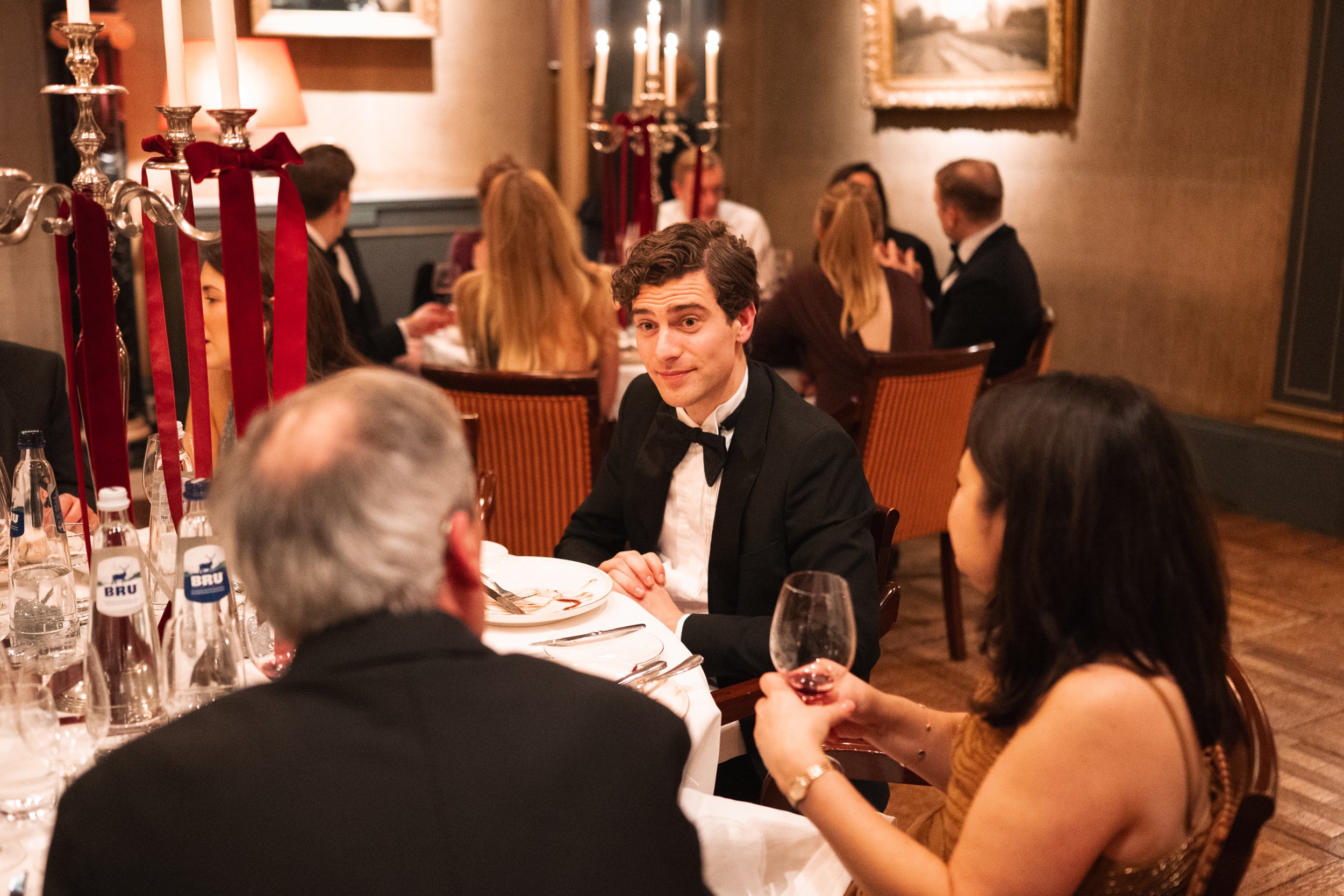 People in formal attire dining at a restaurant, with a man in the center wearing a tuxedo, holding a wine glass, and engaging with others around the table.