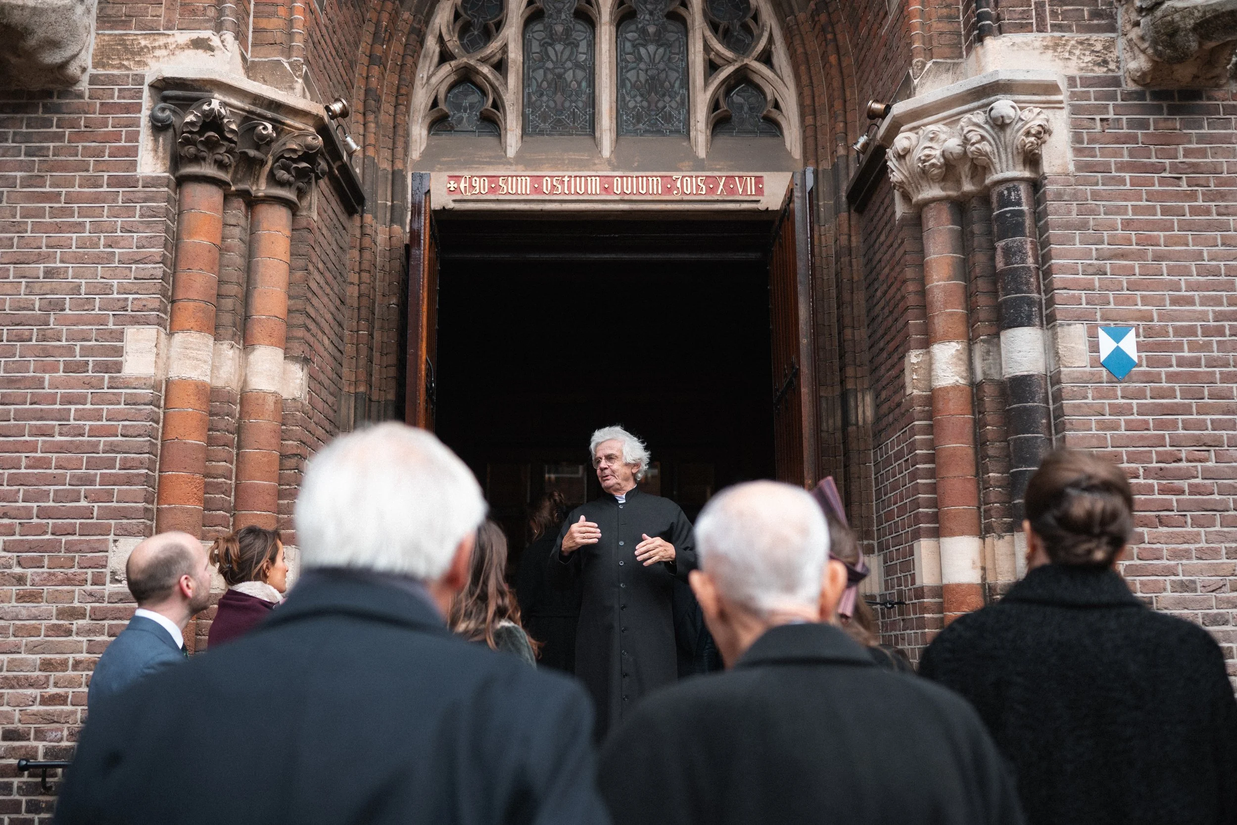 A priest speaking to a group of people outside a brick church with Gothic architecture. The priest is in black clergy robes, and the group appears to be listening attentively.