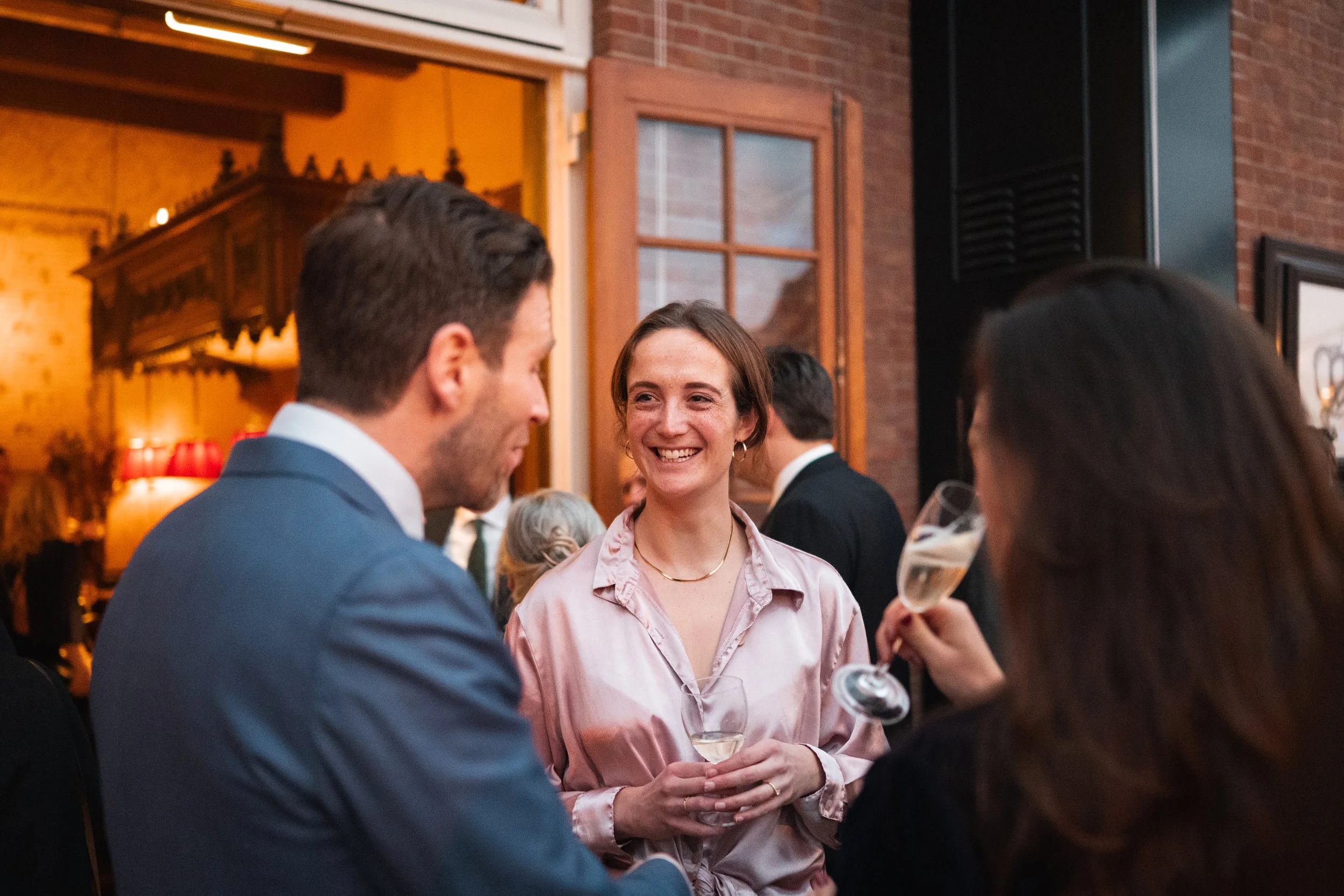 People socializing at a gathering, woman smiling while holding a glass of wine, man in a suit talking, indoor setting with warm lighting and brick walls.