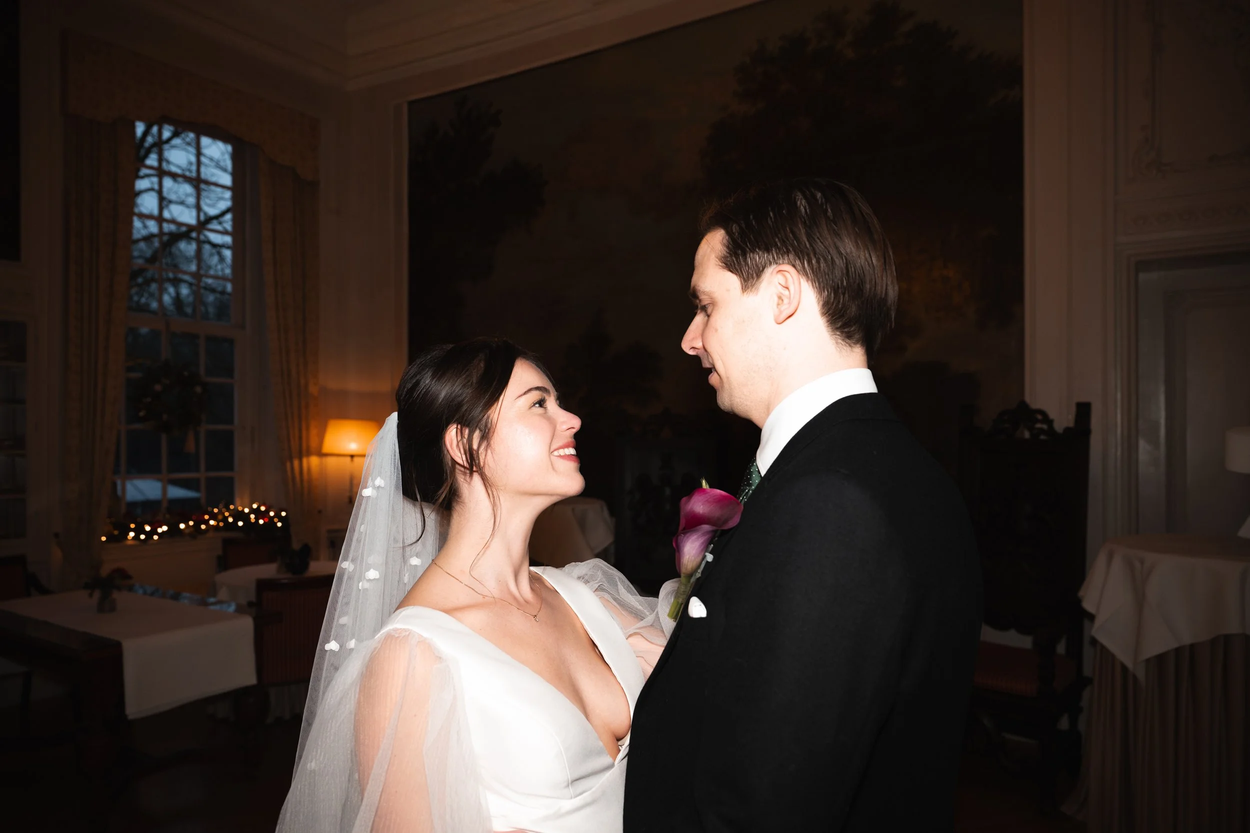 A bride and groom looking at each other indoors at night with warm lighting and decorated windows in the background.