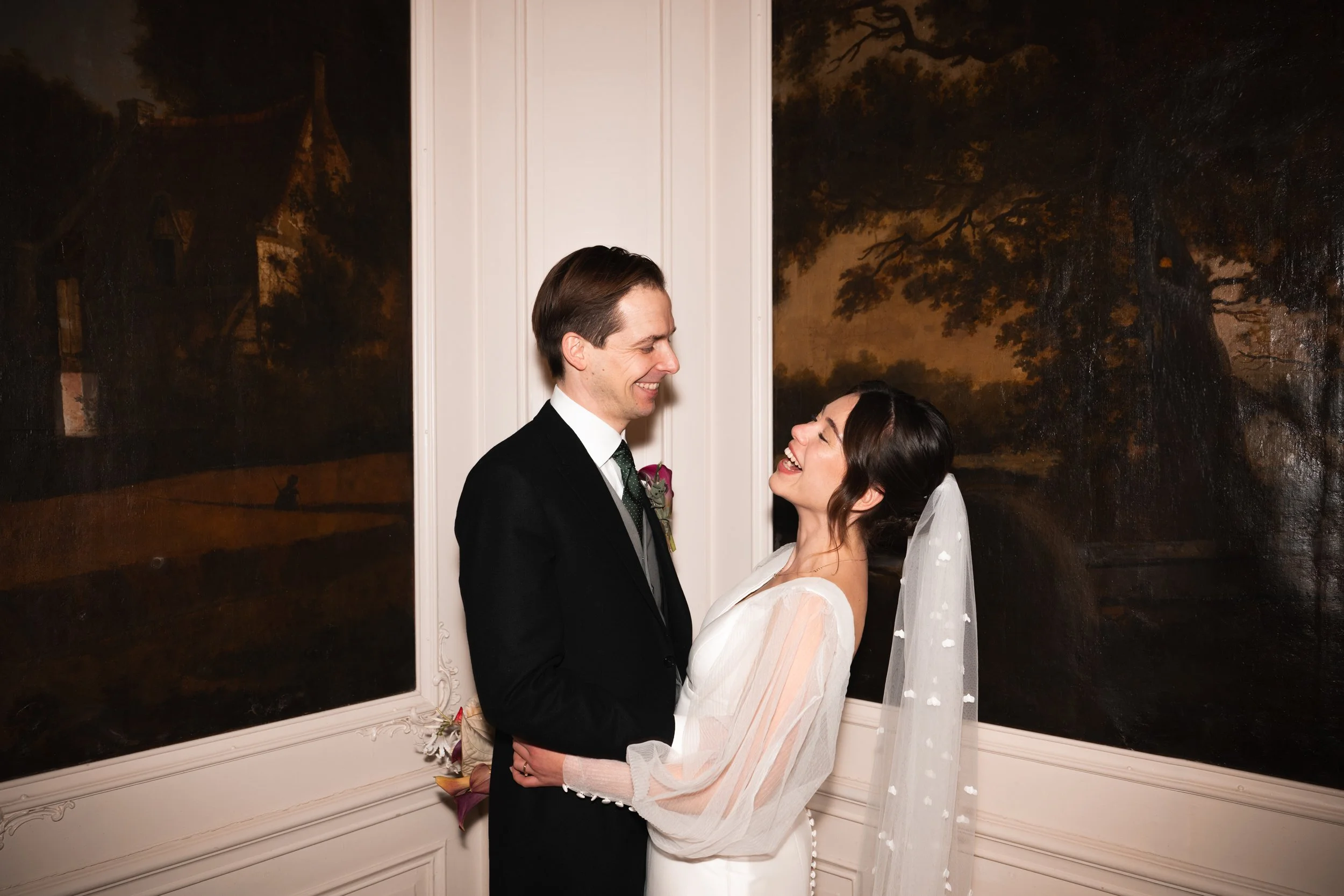 A newlywed couple in wedding attire, smiling and holding each other, standing in front of a wall with dark landscape paintings.