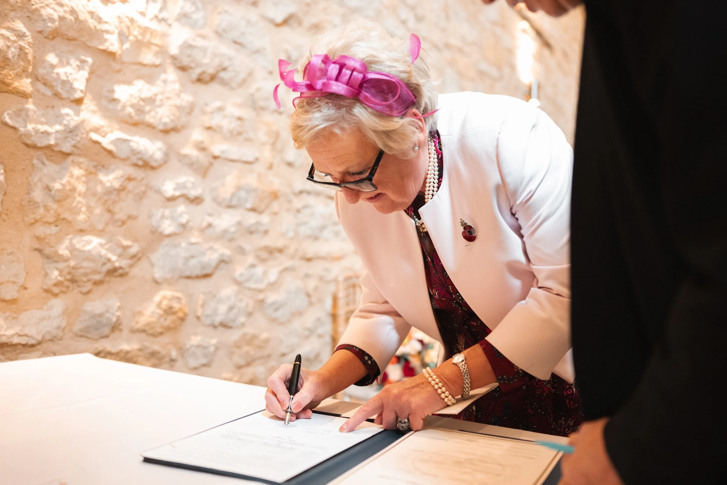An elderly woman with blonde hair, pink ribbon fascinator, glasses, pearl necklace, and jewelry, signing a document on a table with a pen. She is wearing a cream-colored jacket with a brooch and a floral dress.