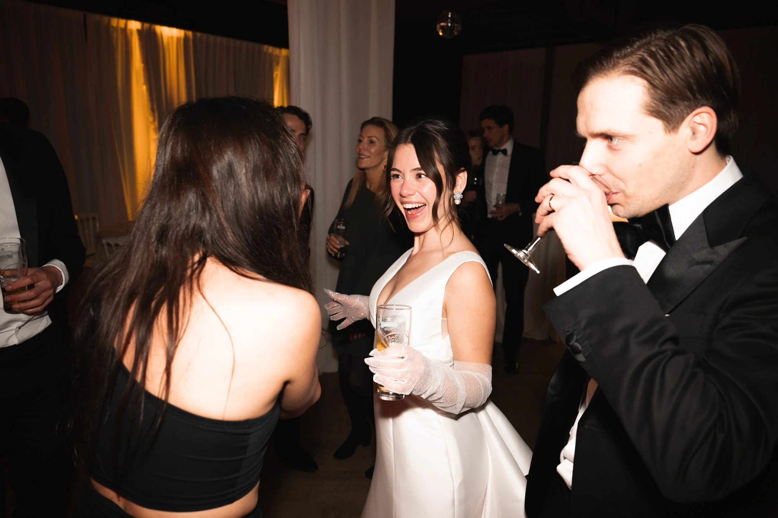 A group of people at a formal event, with a woman in a white dress and gloves smiling and talking to a woman with long dark hair in a black strapless top, while a man in a tuxedo drinks from a glass.