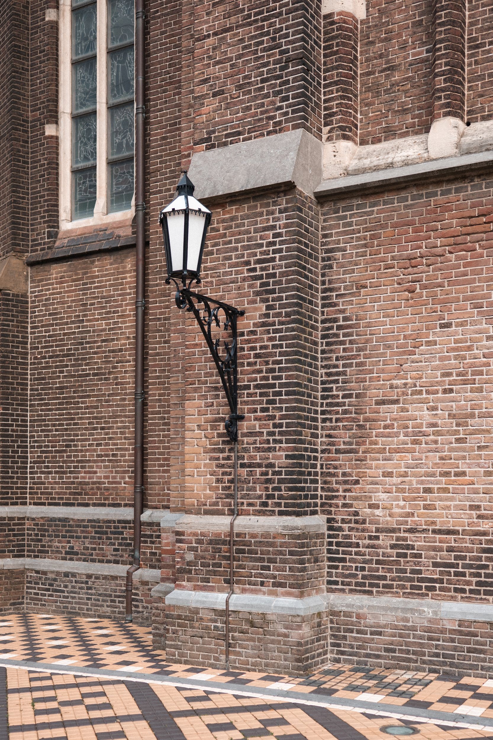 A black, vintage-style street lamp mounted on a brick wall of a building with large windows.