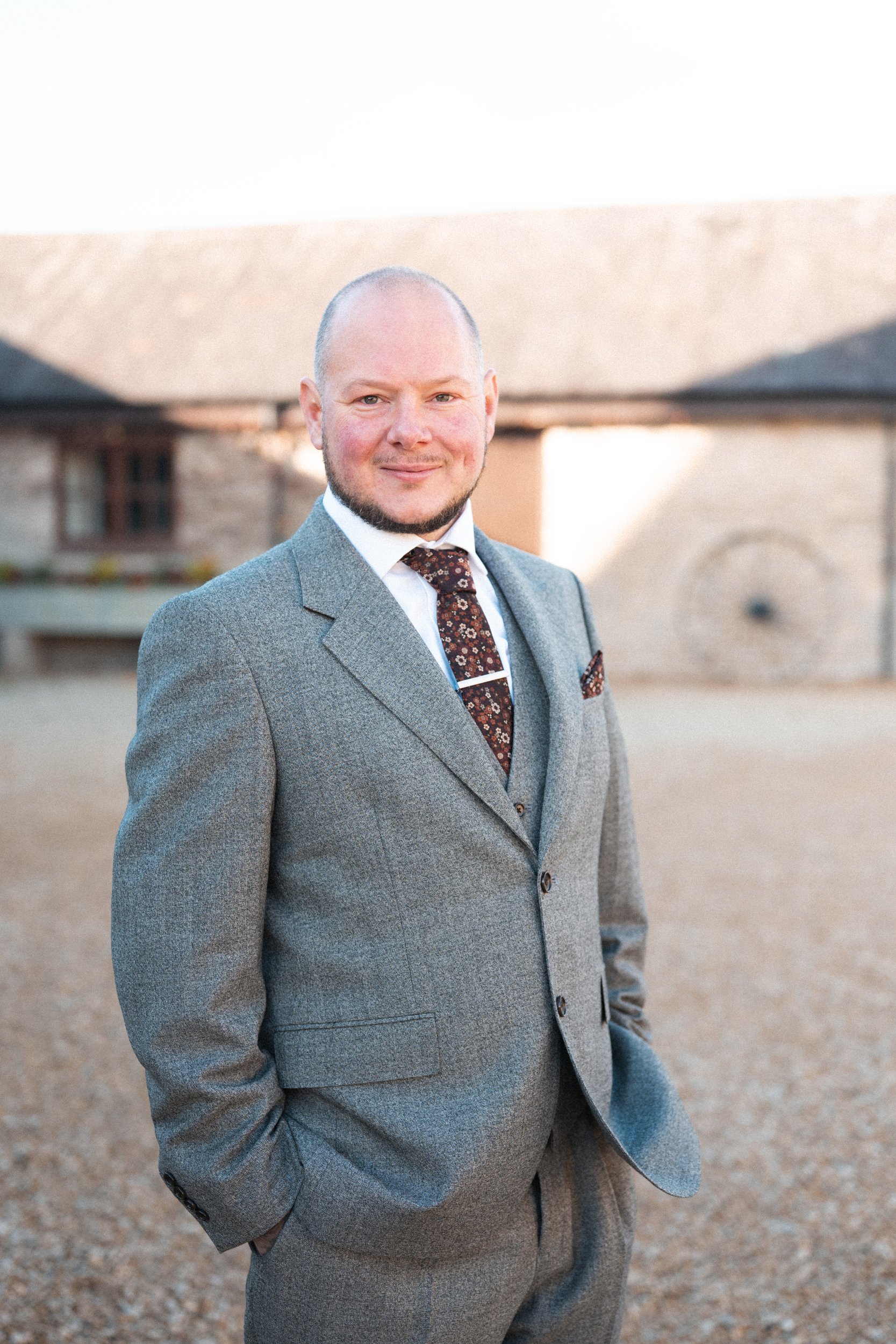 A man in a grey suit, white shirt, and patterned tie stands outdoors with a blurred building in the background.