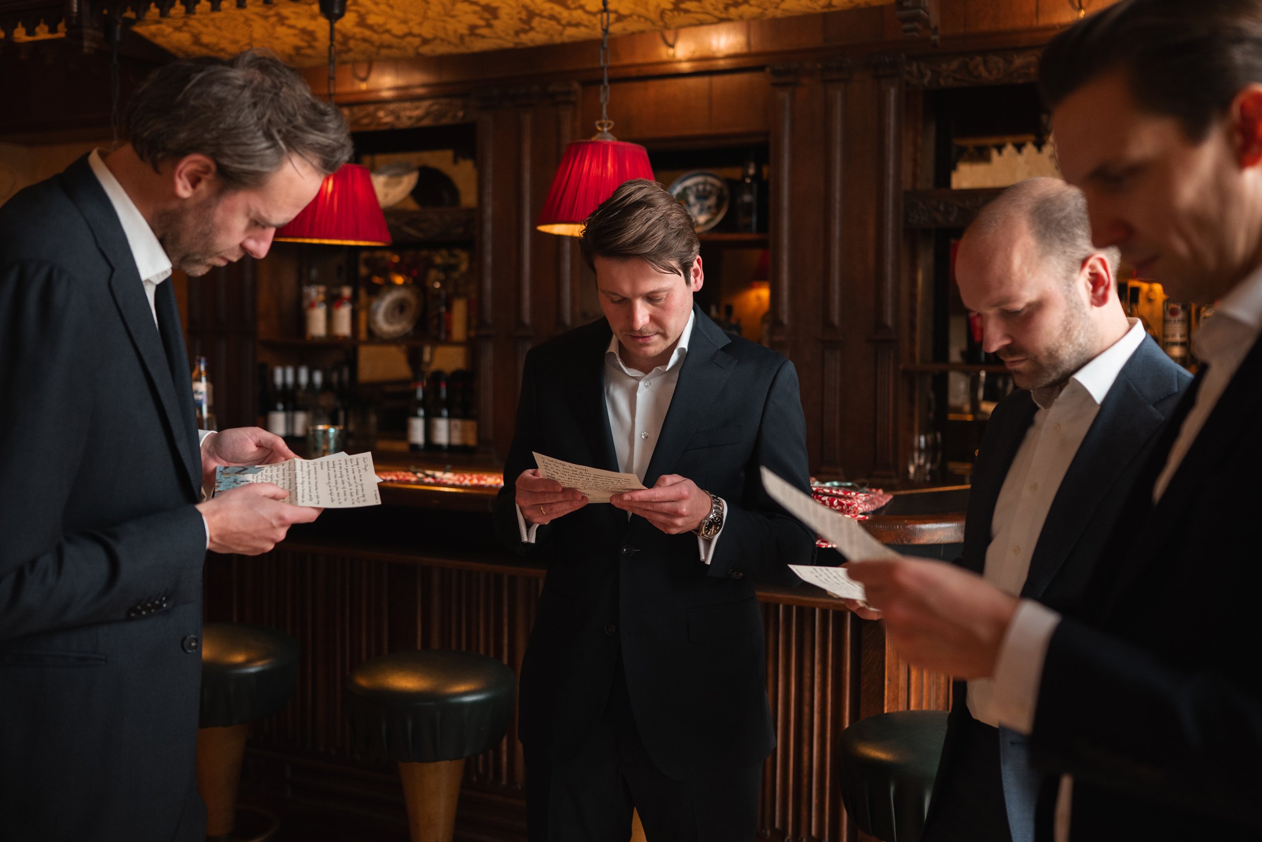 Four men in suits standing at a bar, reading menus in a dimly lit, wood-paneled bar with red lampshades.