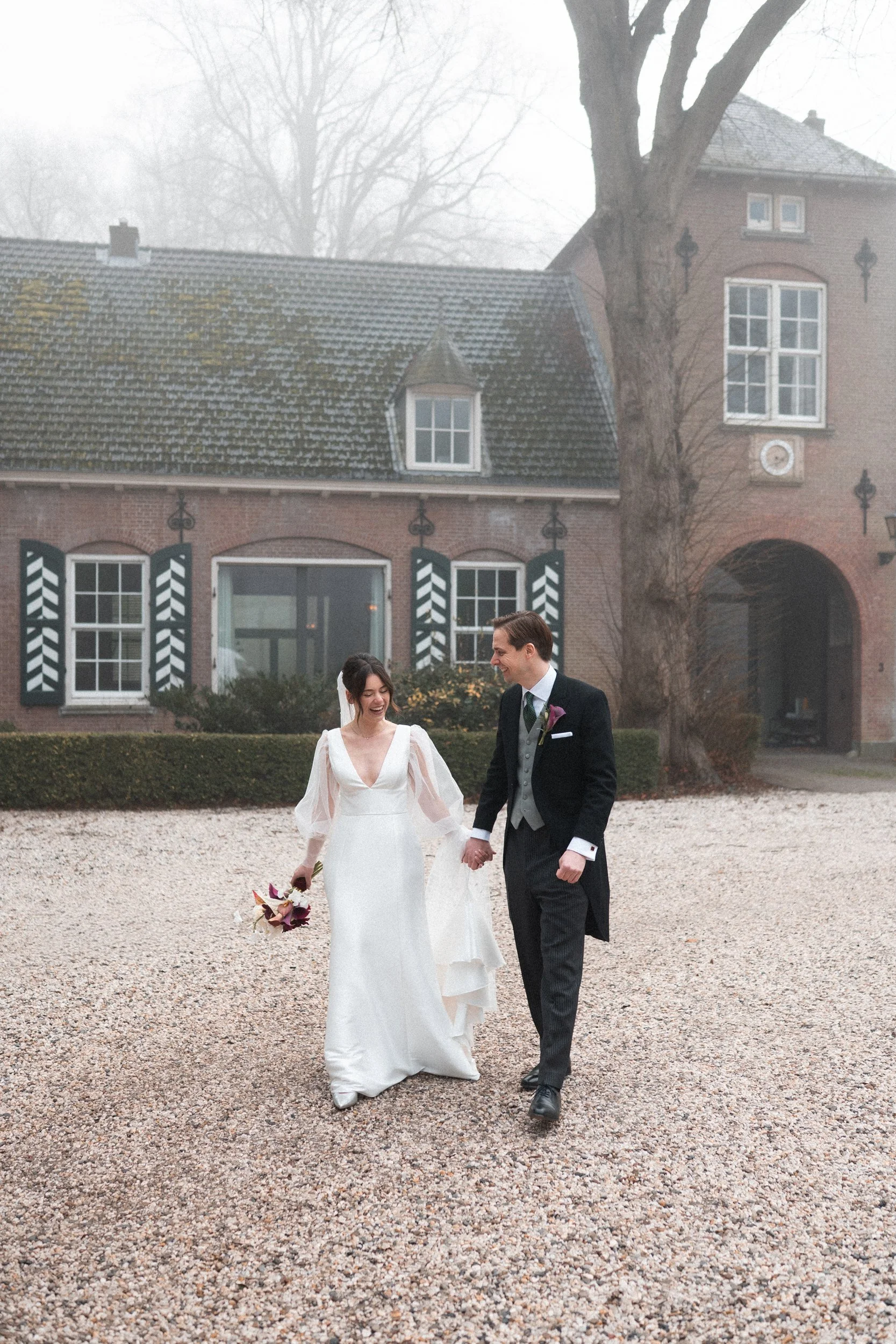 A newlywed couple walking hand in hand outside on a gravel path, smiling and dressed in wedding attire, with an old brick building and leafless trees in the background on a foggy day.