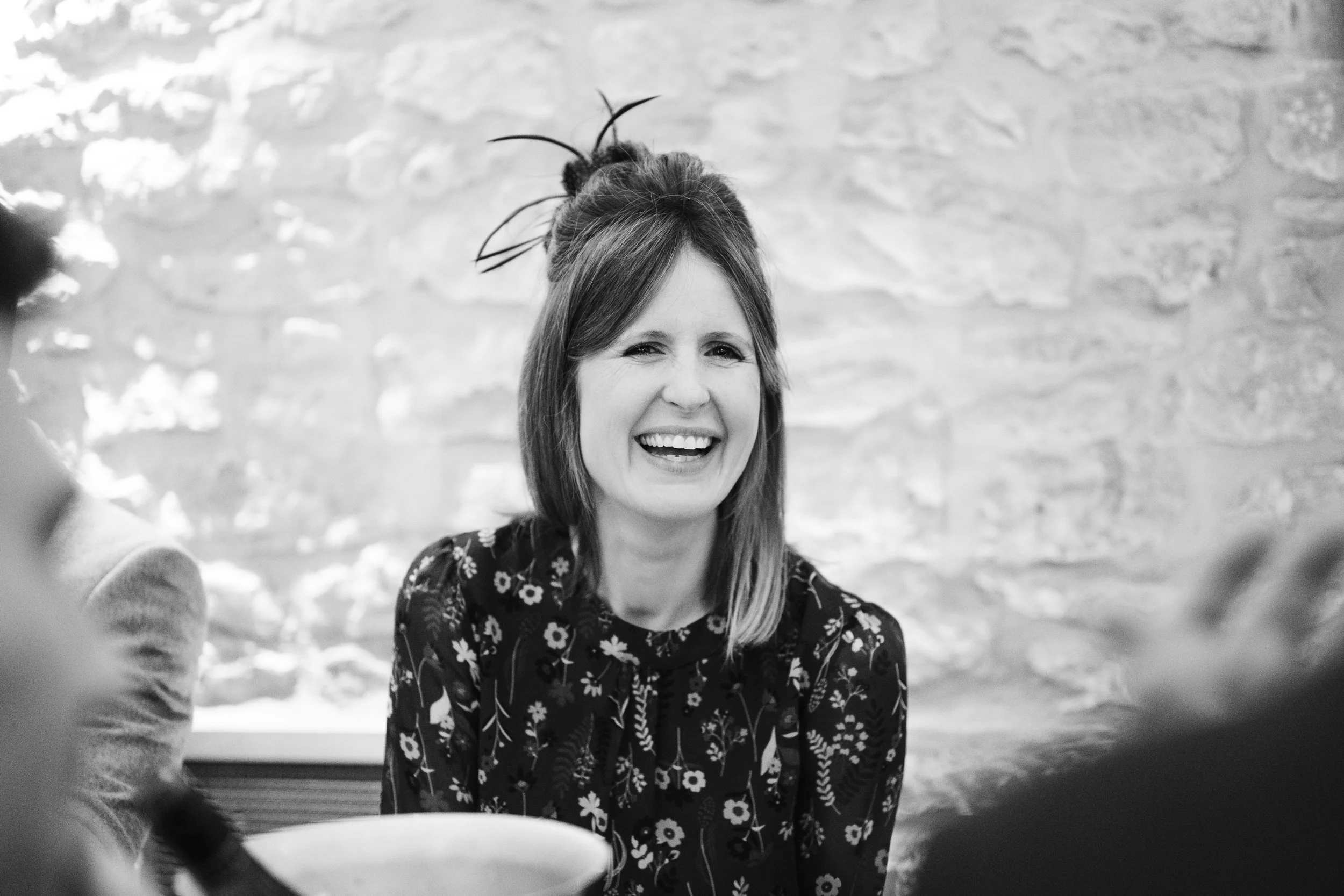 A woman with shoulder-length hair and a floral dress, smiling and winking, sitting in front of a brick wall.