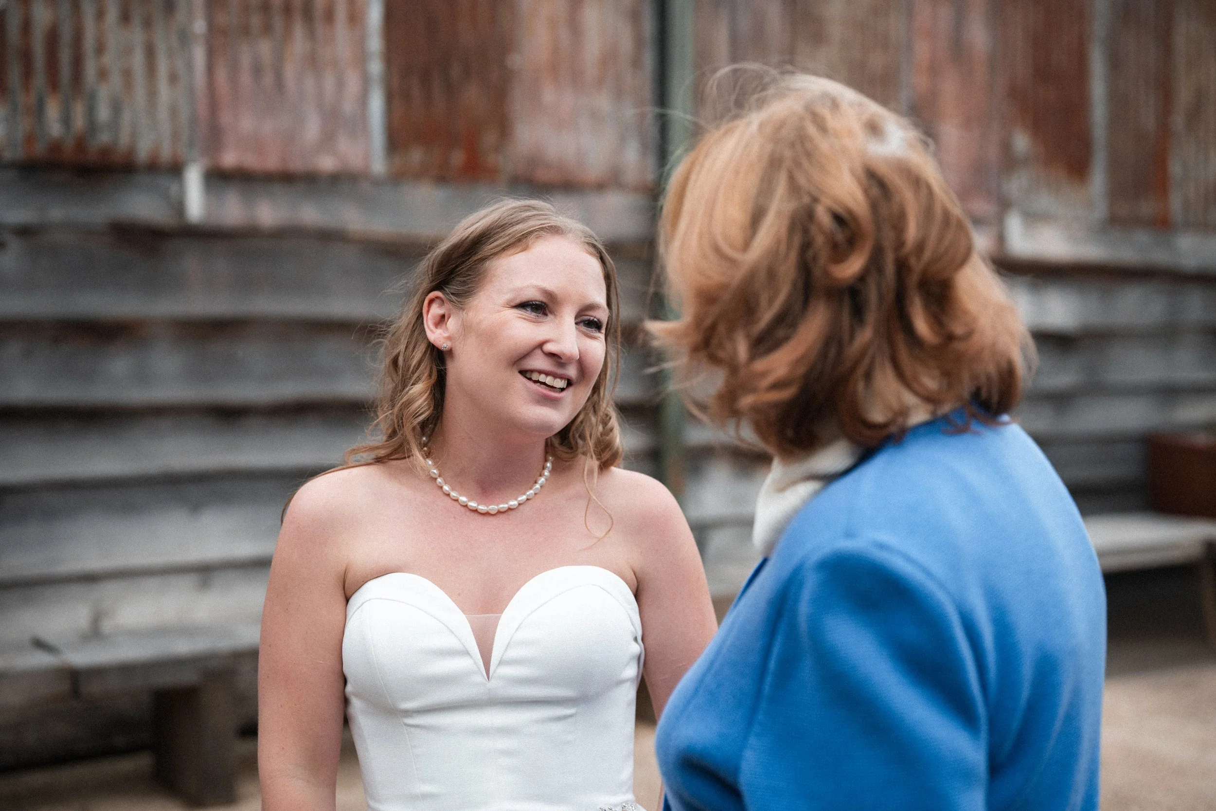 A happy woman in a white strapless wedding dress and pearl necklace smiling and talking to another woman with red hair wearing a blue jacket.