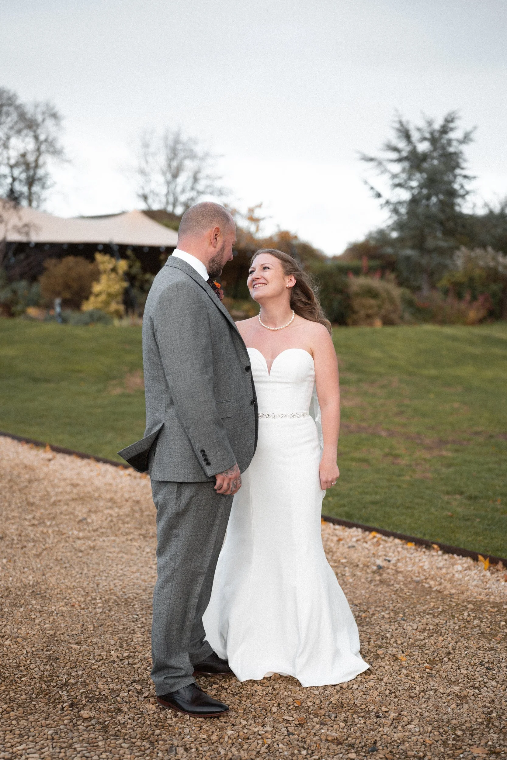 A bride and groom standing outdoors on a gravel path, facing each other and smiling, with trees and a tent in the background.