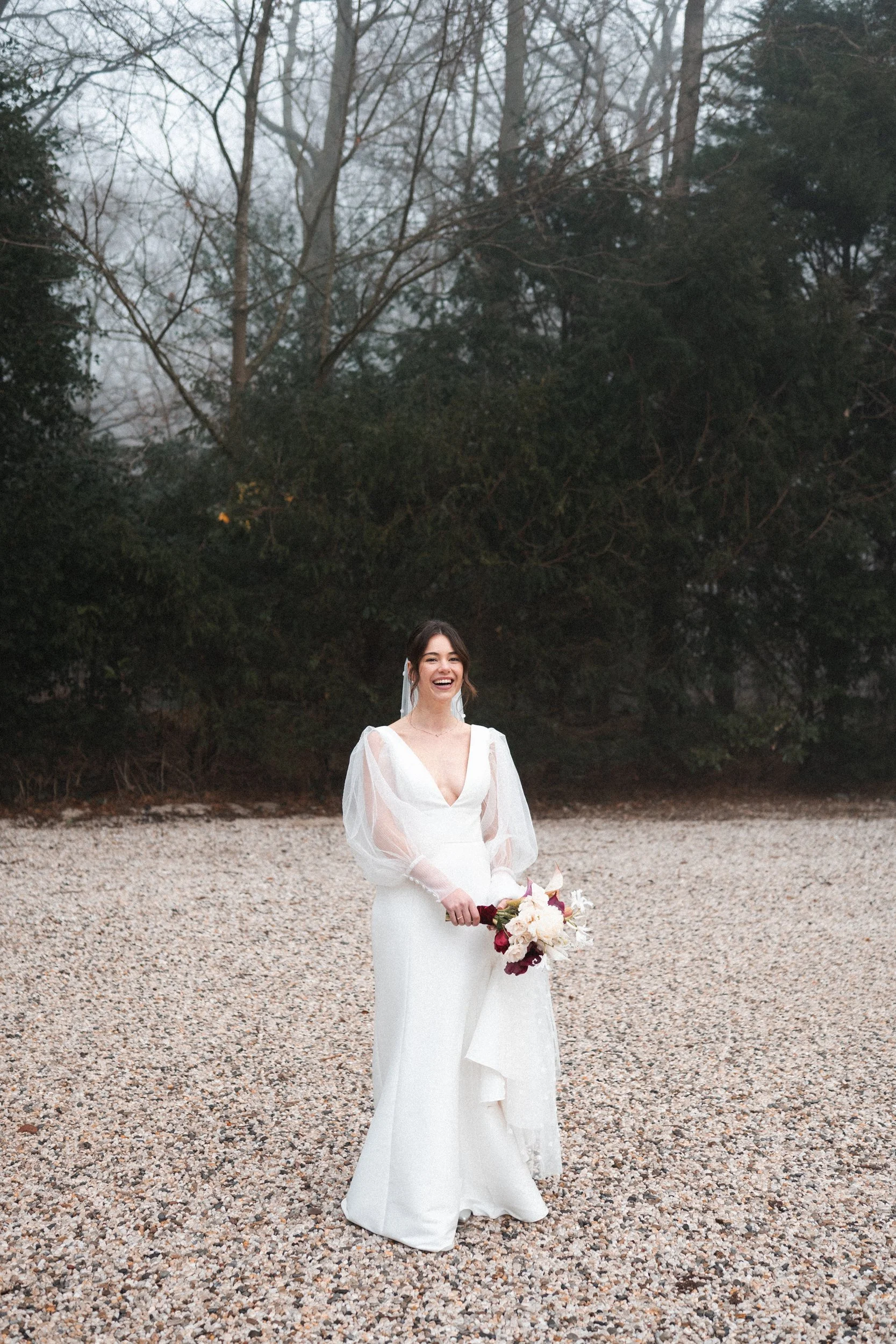 A bride in a white wedding dress with sheer puff sleeves, holding a bouquet of flowers, standing on a gravel surface outdoors with trees and bushes in the background, smiling.