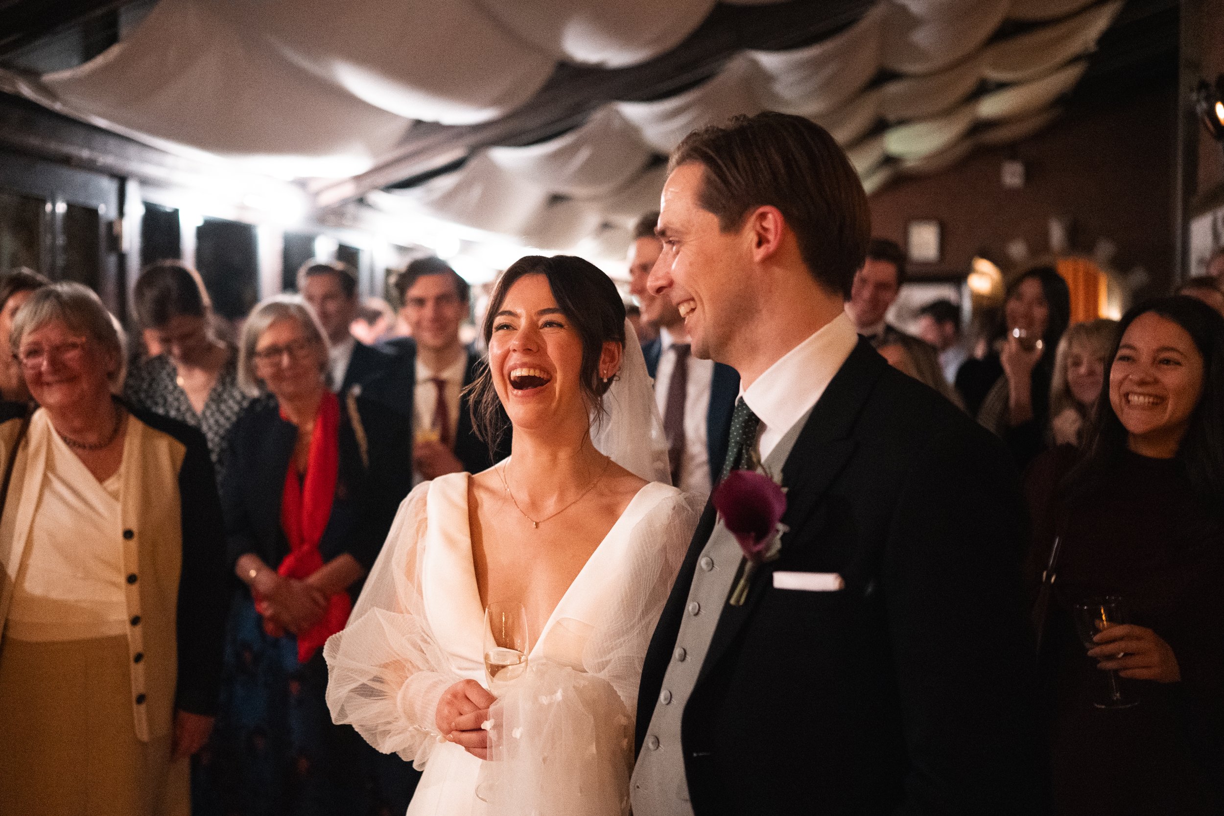 A bride and groom at a wedding reception, smiling and laughing, surrounded by guests.