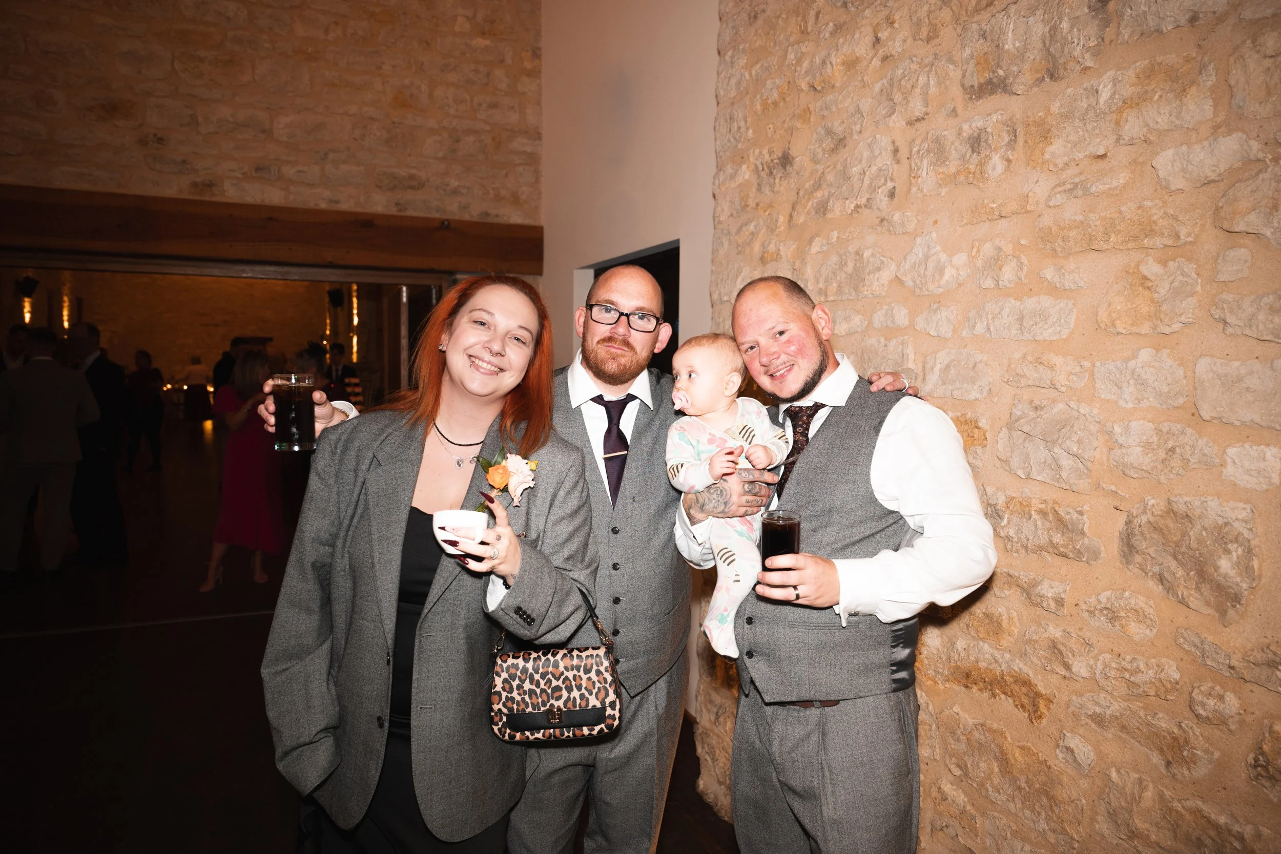 Group of four adults and one baby at a social event, standing against a stone wall, holding drinks and smiling at the camera.