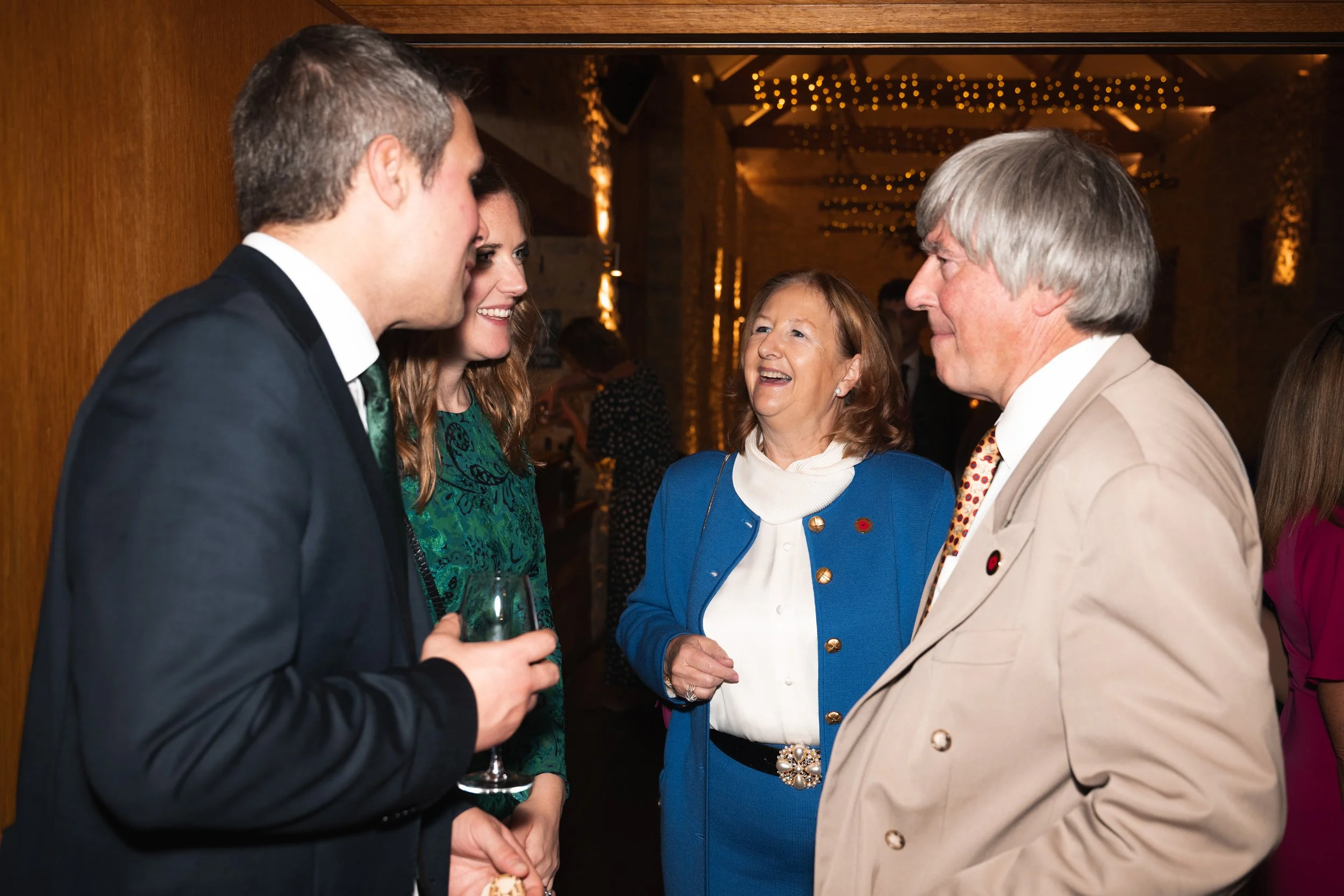 Four people in conversation at a social event, smiling, with a warm indoor setting and string lights in the background.