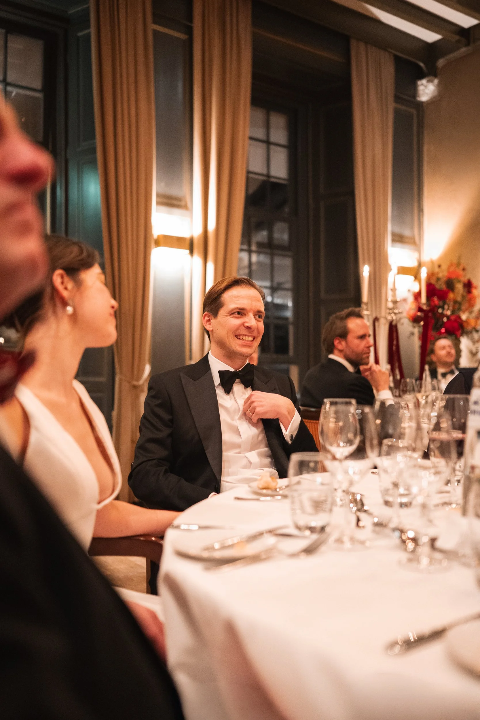 A man in a tuxedo smiling at a formal dinner event, seated at a table with wine glasses and a woman in a white dress in a sophisticated, beautifully decorated room with curtains and floral arrangements.