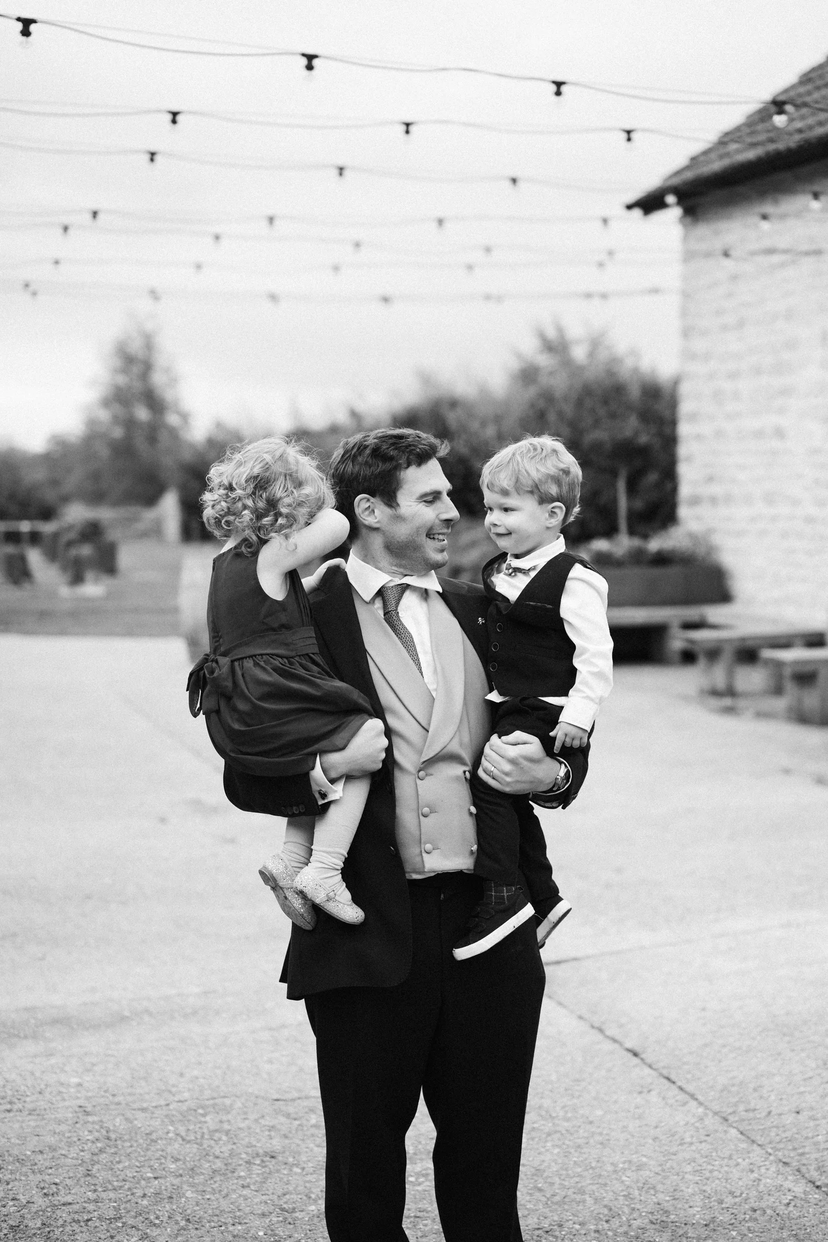 A man in formal attire holding two young children, one girl and one boy, outdoors with string lights overhead and a building to the side, black and white photograph.
