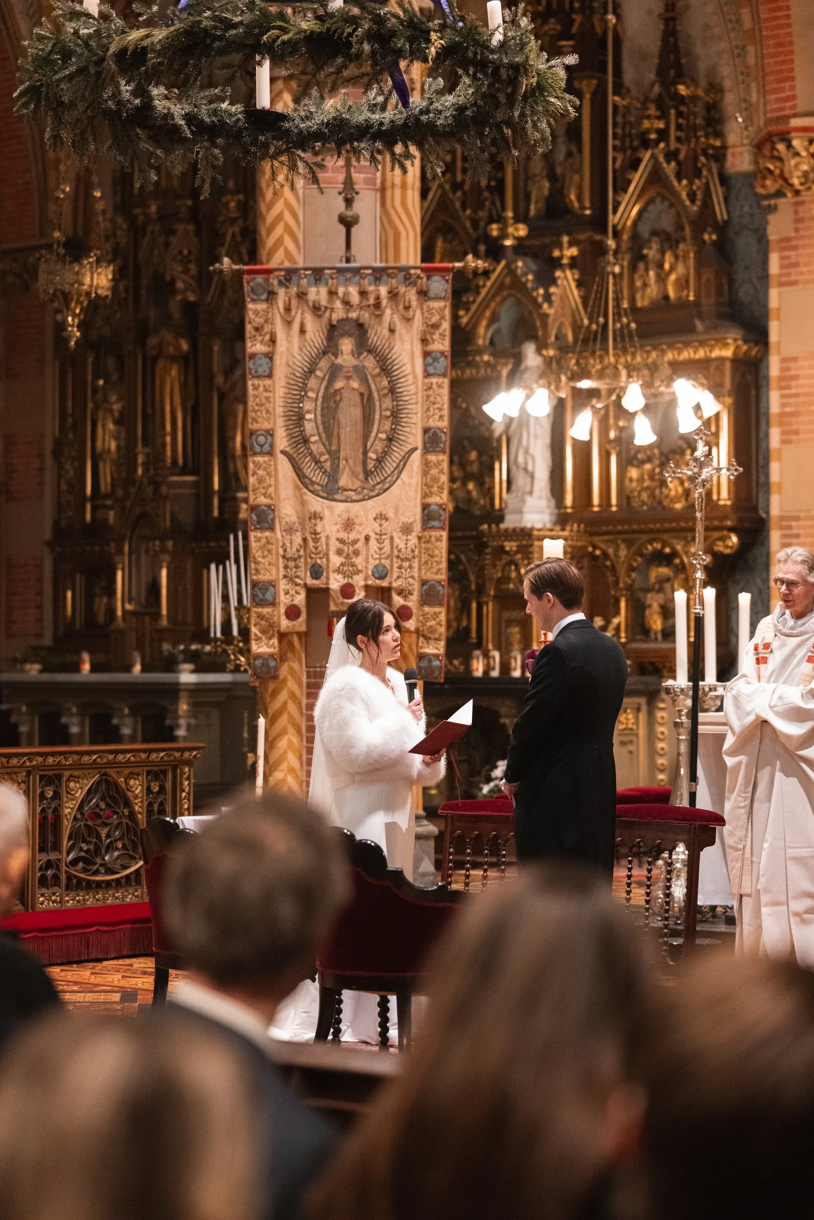 A couple getting married during a church ceremony, with a priest present. The bride is holding a microphone and a paper, wearing a white fur shawl, and the groom is dressed in a black suit. The church interior features ornate gold decorations, candle