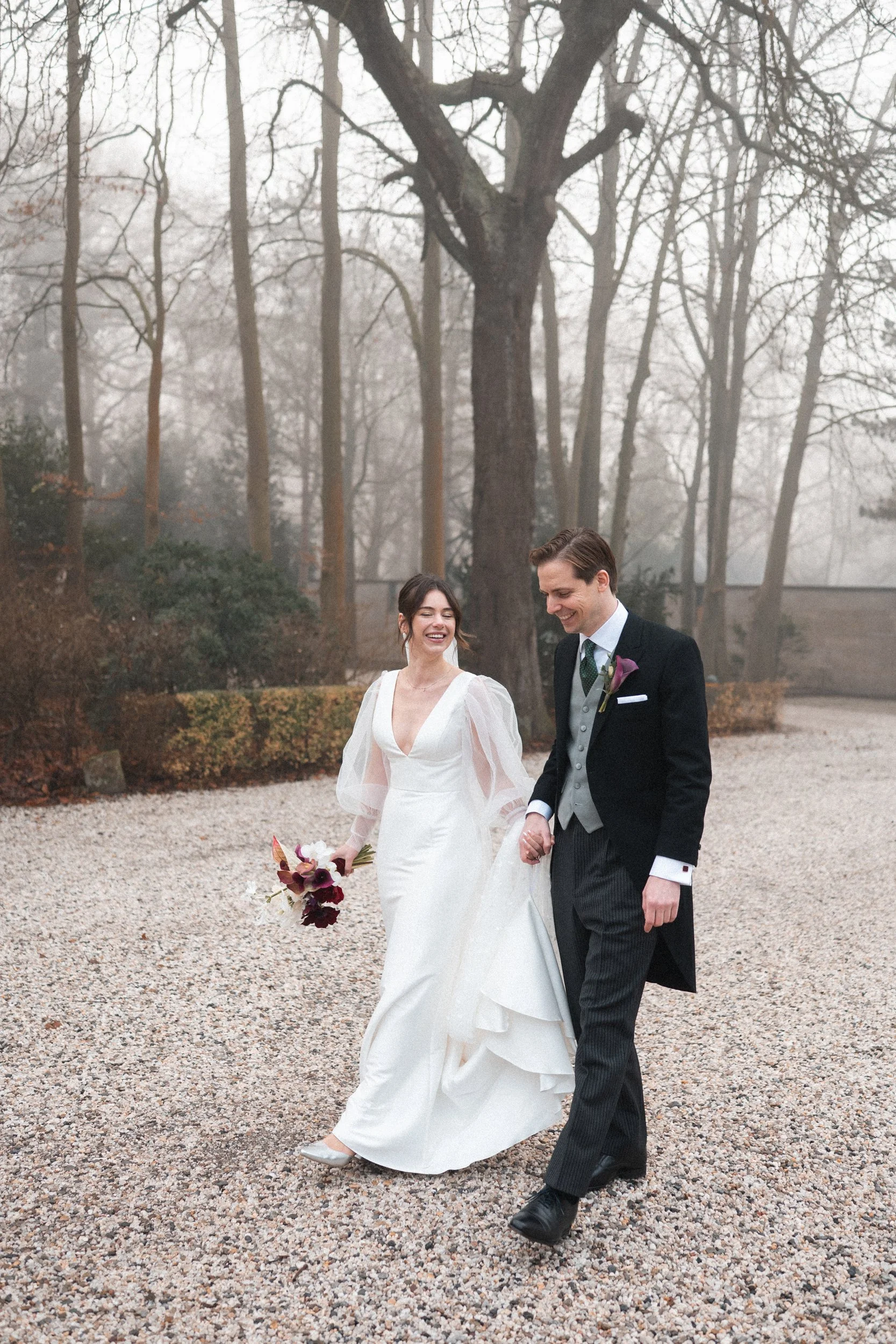 A bride and groom walking outdoors on a cloudy day, smiling and holding hands, with trees and shrubs in the background.