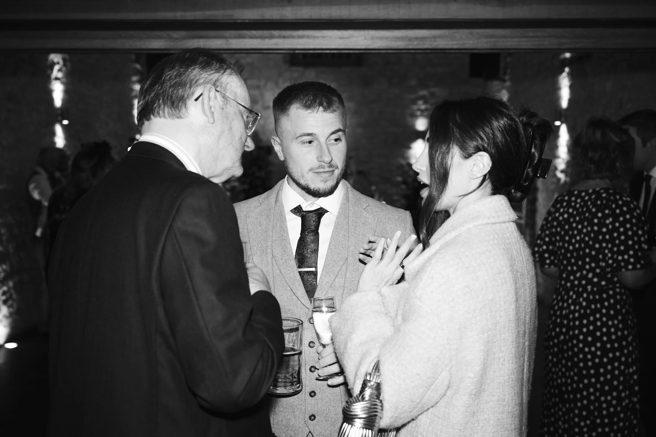 Three people engaged in conversation at a social gathering with other guests in the background.
