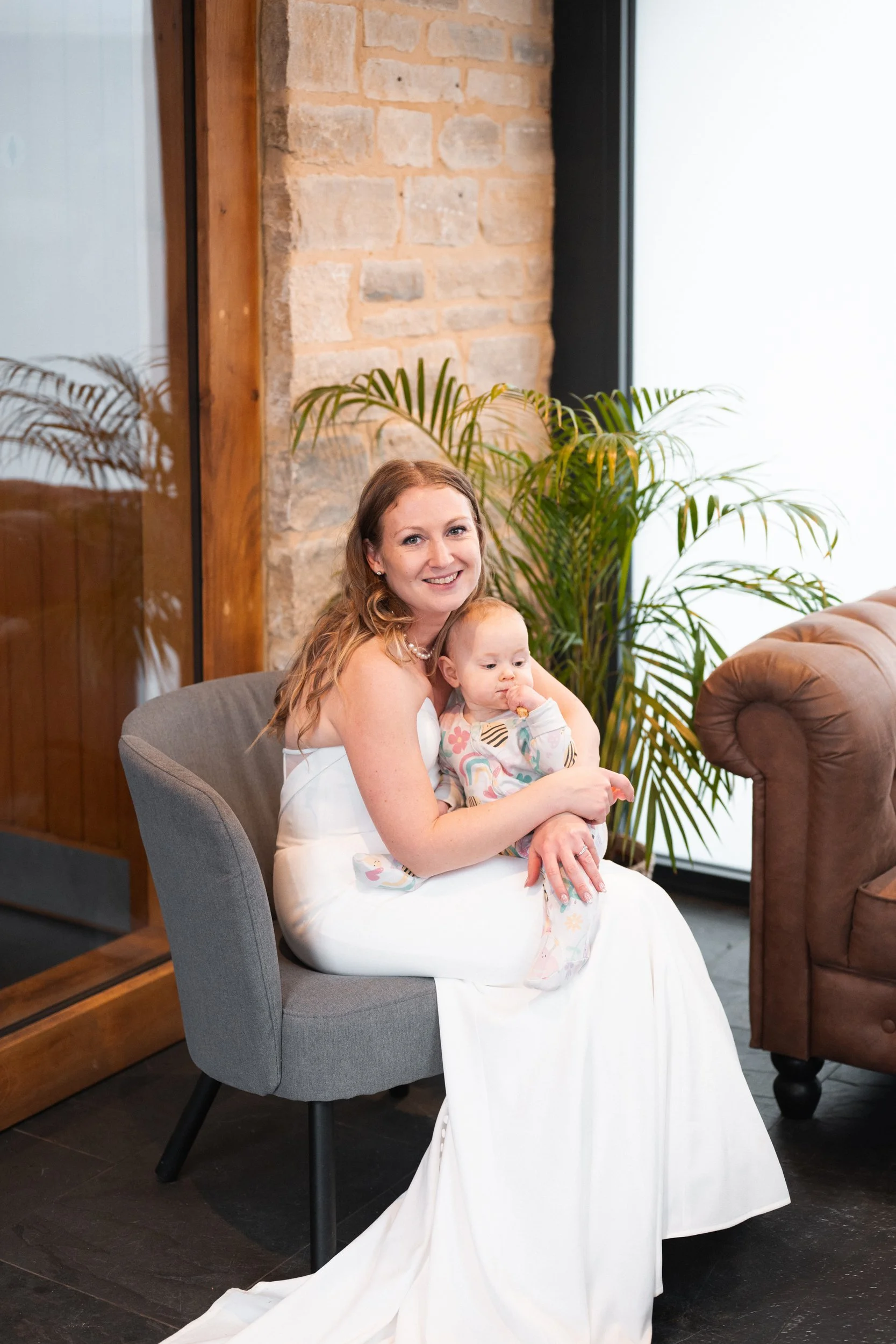 A woman in a white dress is sitting on a gray chair holding a baby, both smiling. They are indoors with a brick wall, a large window, and green plants in the background.