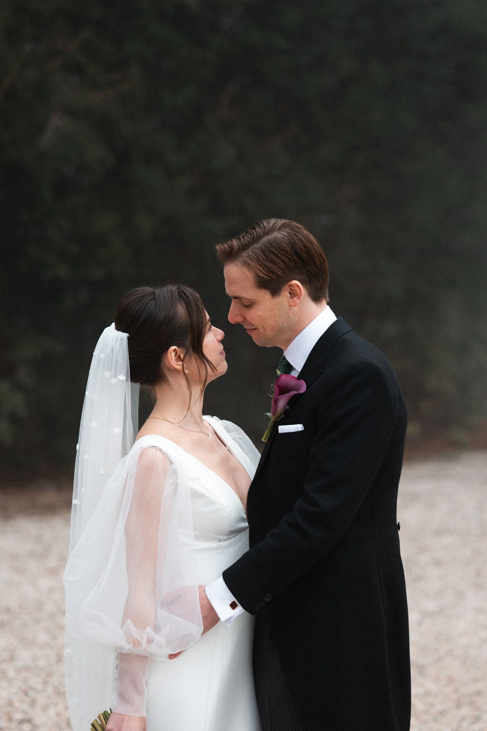 Bride and groom standing close together outdoors, gazing into each other's eyes, holding hands, with a foggy or misty background.