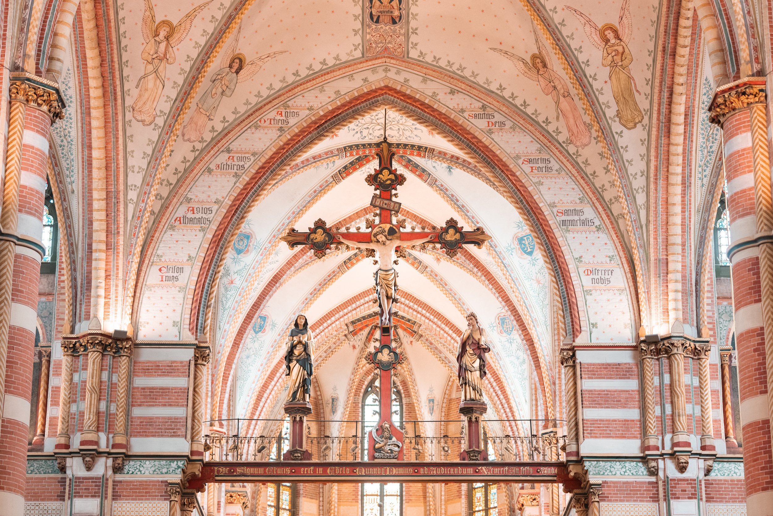 Interior of a church with a high vaulted ceiling, depicting a crucifix with Jesus Christ in the center, flanked by statues of saints, and surrounded by Latin inscriptions and angel murals.