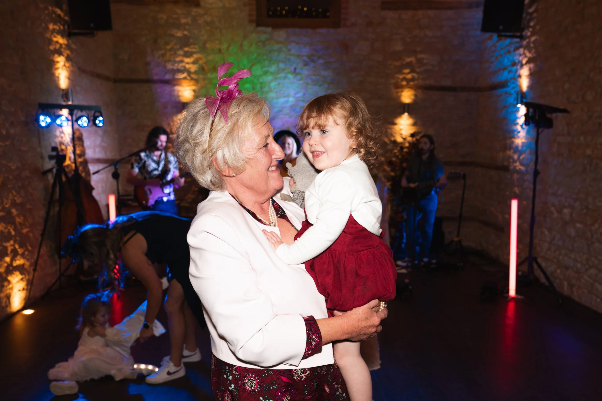 An elderly woman joyfully looking at a young girl in a party setting, with a band playing in the background and children dancing.