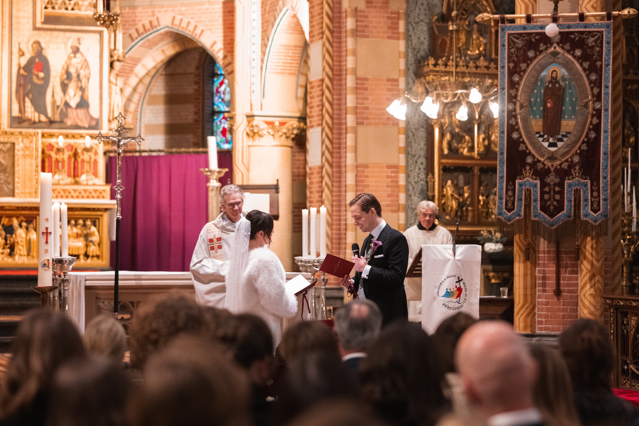 A wedding ceremony taking place inside a church with clergy and a couple. The priest is standing next to a bride and groom, who are facing each other. The officiant is holding a book, and the couple is holding hands. The church features ornate decora