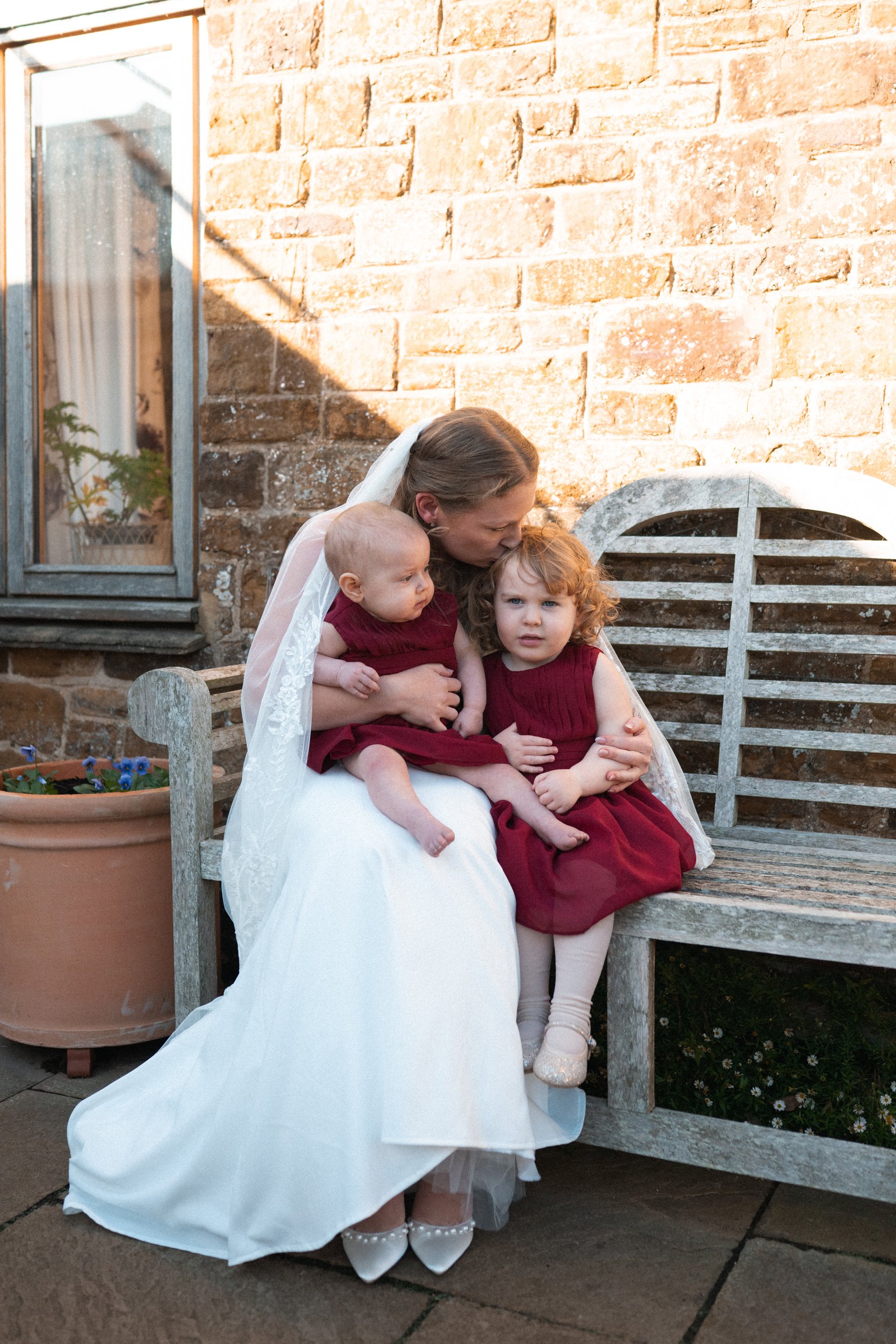A woman dressed as a bride is sitting on a wooden bench with two young girls, all wearing burgundy dresses, outside near a brick wall. The woman is kissing one girl on the head, and the other girl looks directly at the camera. The scene is bathed in 