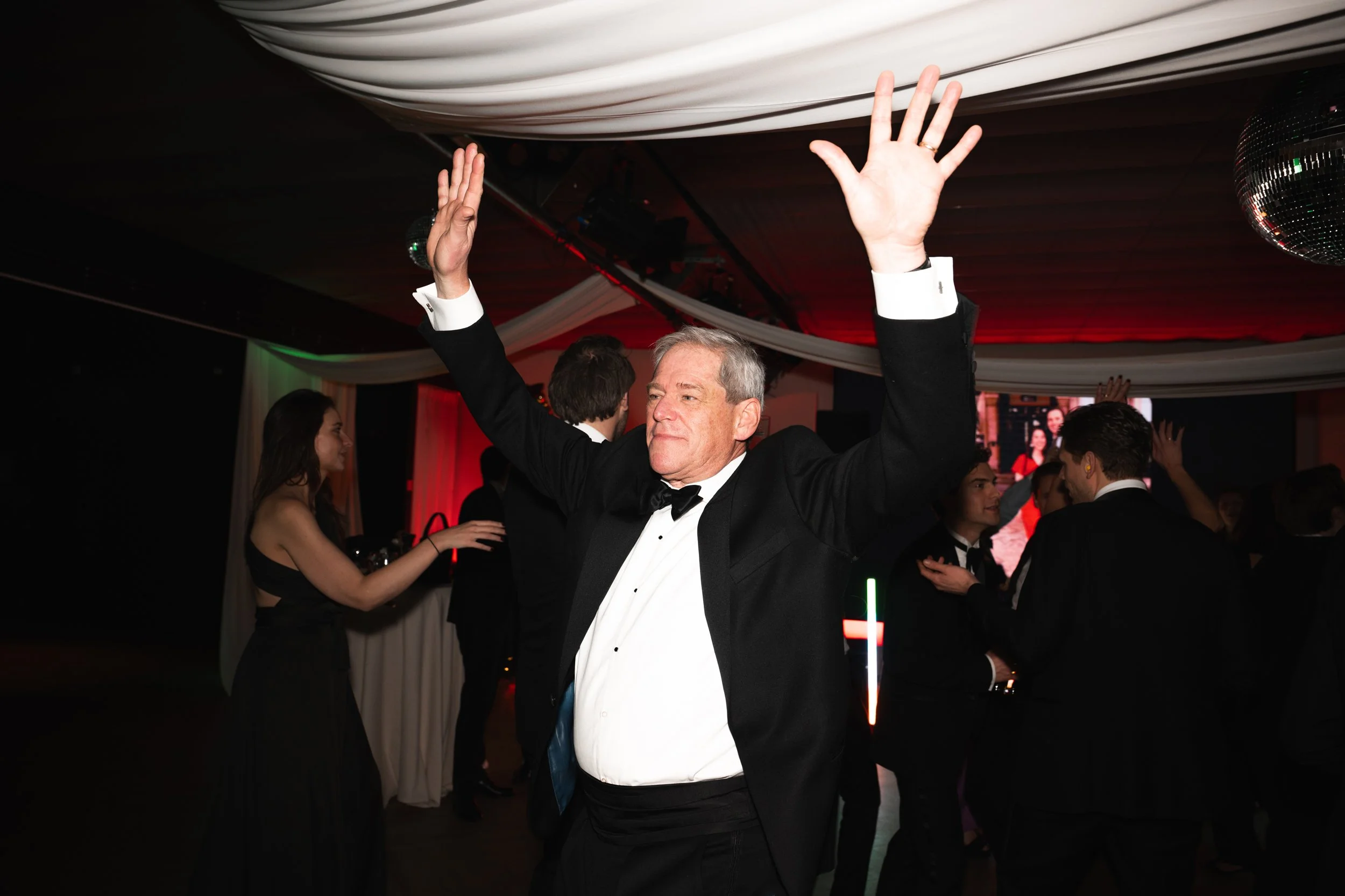 Older man in a tuxedo dancing with his arms raised at a formal event with other guests in the background.