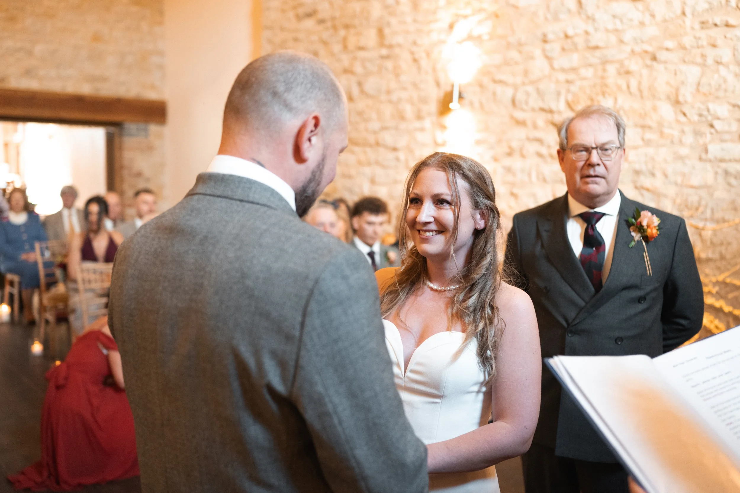 Bride and groom exchanging vows at wedding ceremony with officiant and wedding guests in background.