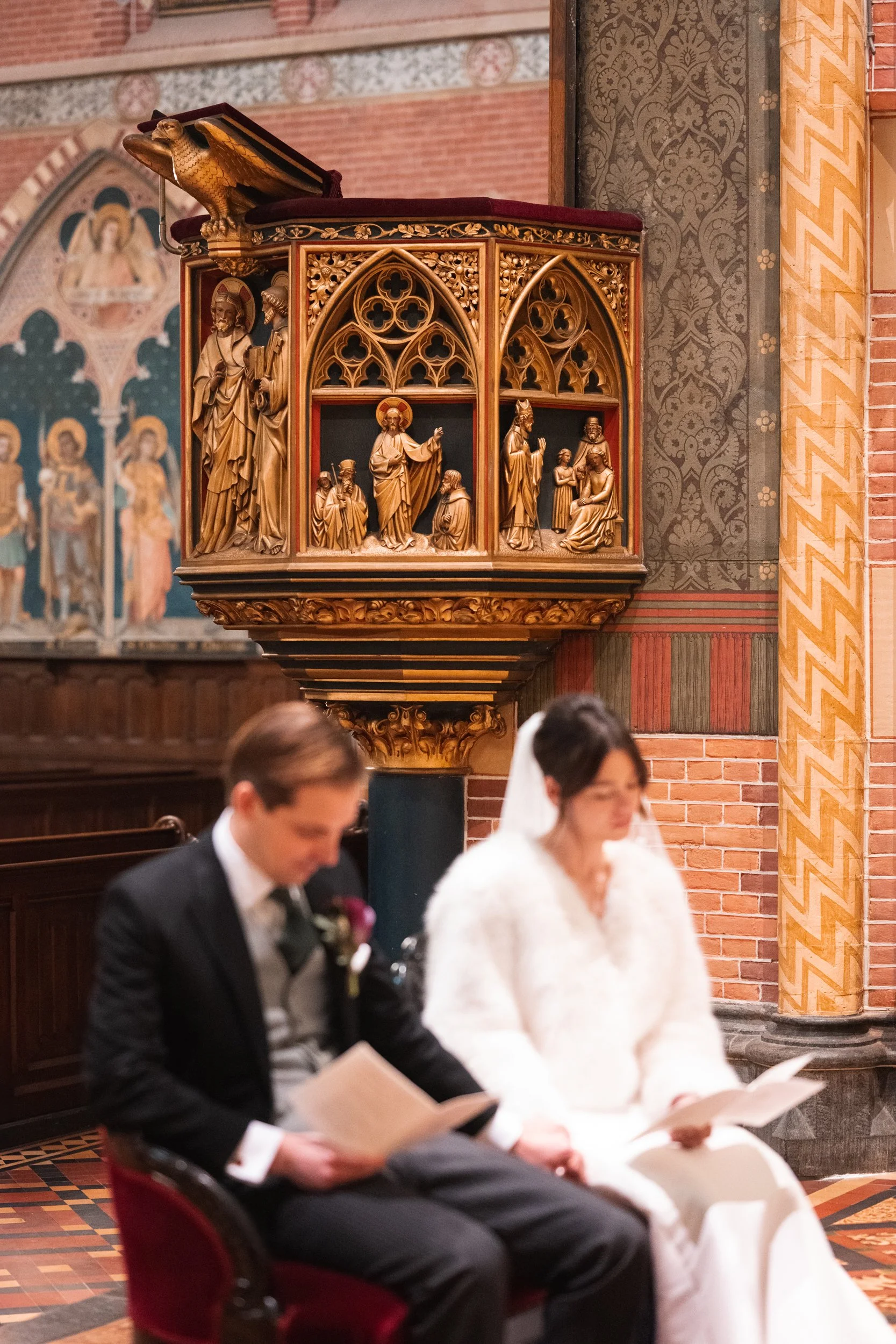 A church interior with a wooden altar featuring religious carvings, and a bride and groom sitting in the foreground, reading from papers during a wedding ceremony.