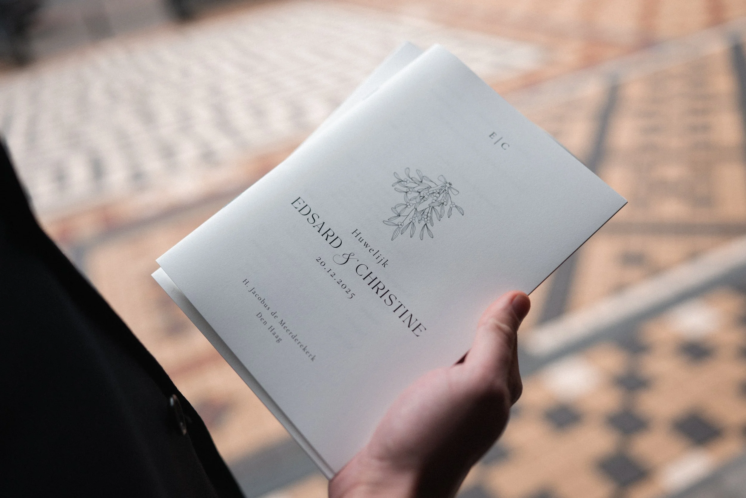 A person holding a wedding invitation with a floral illustration, text in Dutch and English, and a date of December 20, 2025, against a blurred outdoor background.