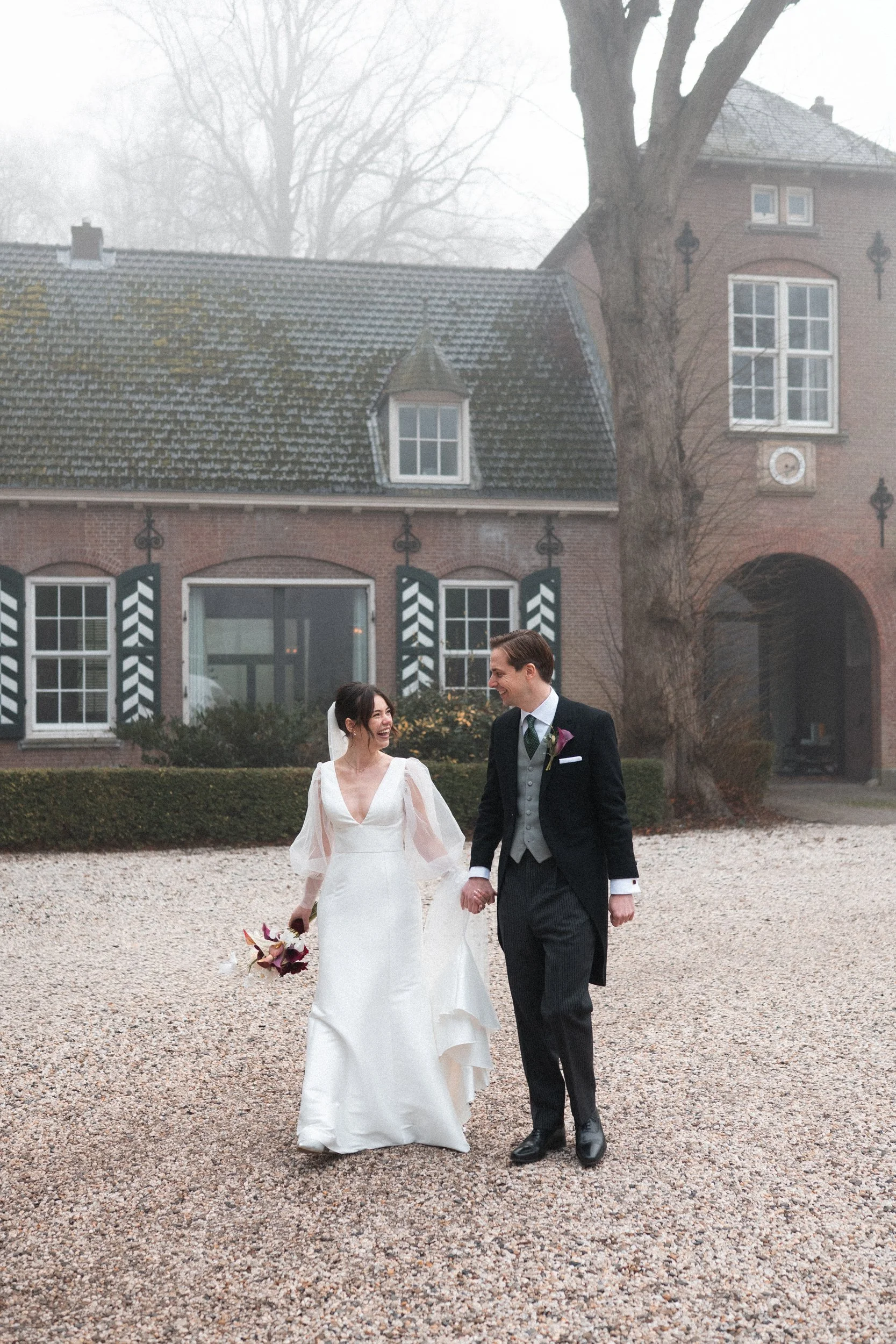 A bride and groom walking hand in hand on a gravel path outside a large brick house on a foggy day, with the bride wearing a white dress and holding a bouquet, and the groom in a black suit with a boutonniere, smiling at each other.