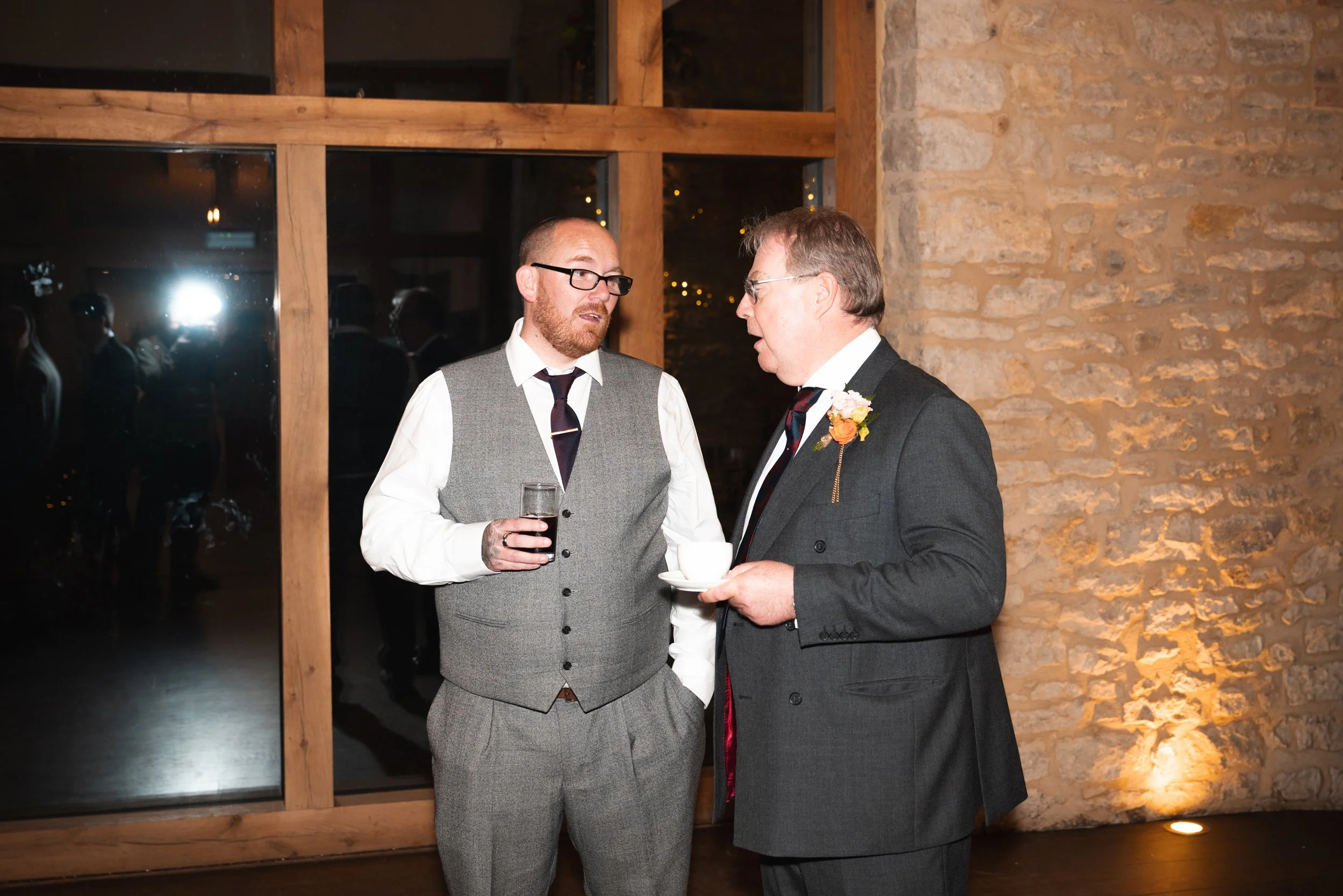 Two men in formal attire having a conversation at an indoor event, one holding a drink and the other holding a teacup, with a brick wall and large window in the background.