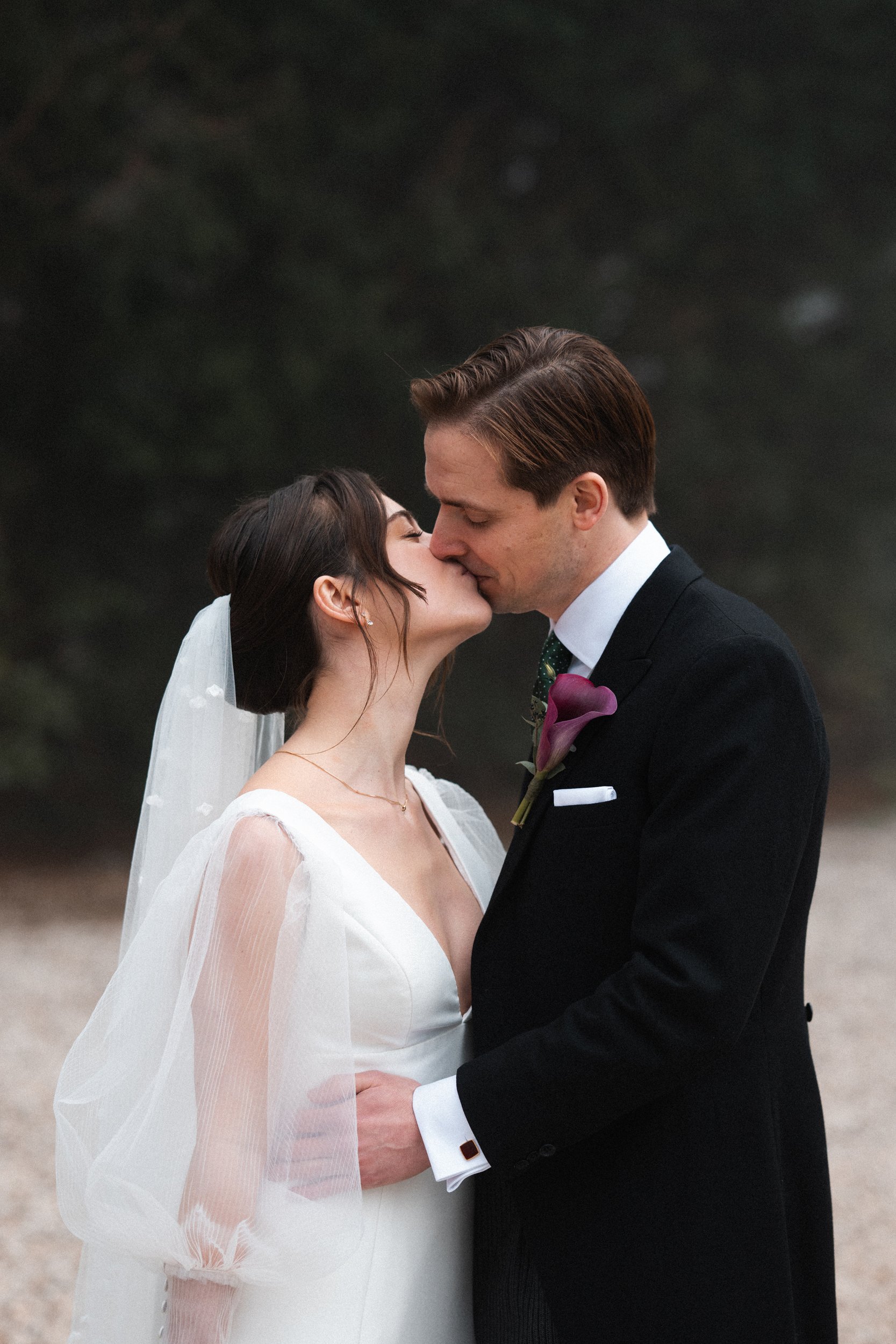 A bride and groom kissing outdoors, the bride wearing a white wedding dress with sheer sleeves and a veil, and the groom dressed in a black suit with a white shirt and green tie, in a natural setting with blurred trees in the background.