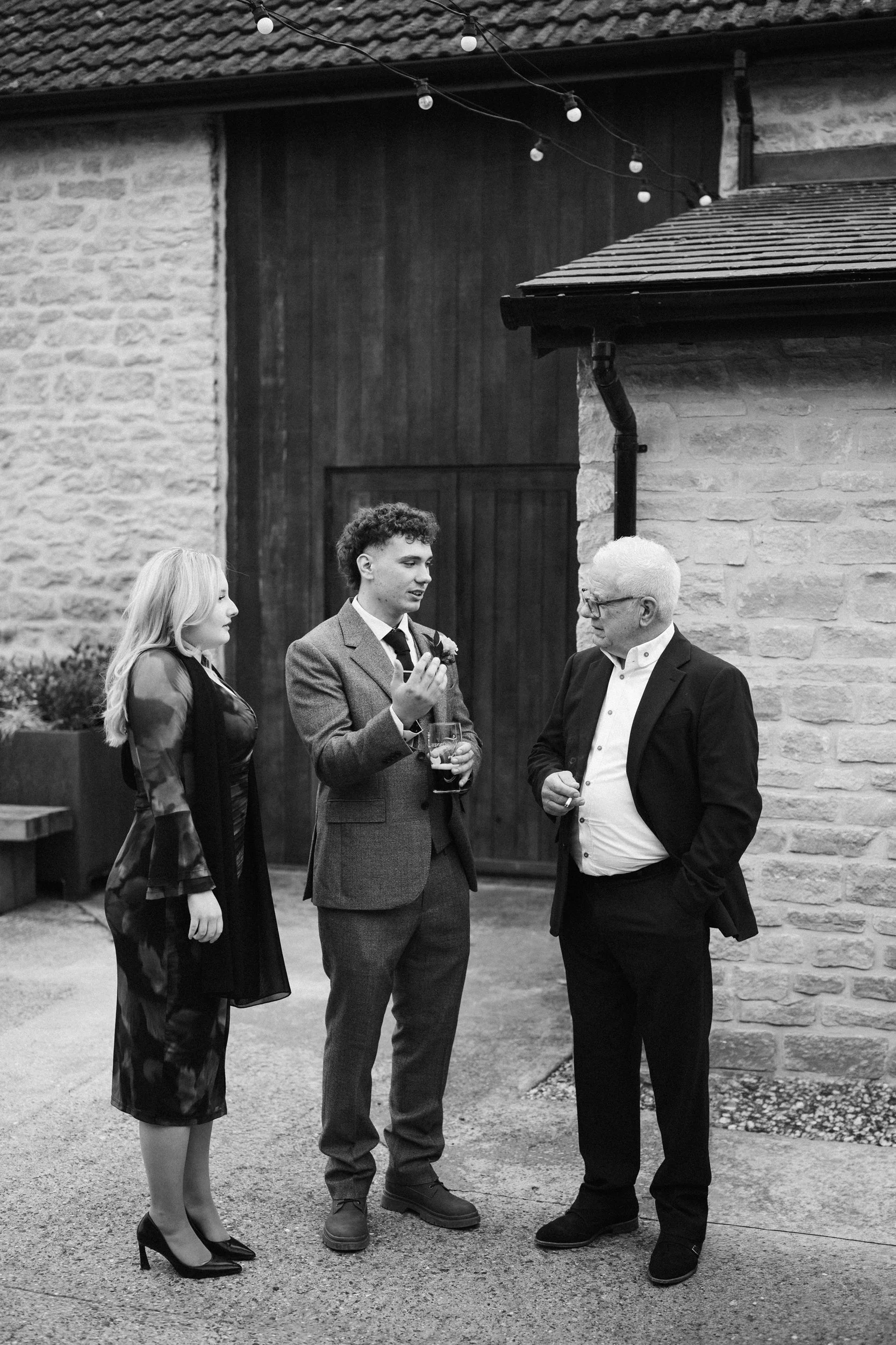 Three people are standing outside near a brick and wooden building, engaging in conversation. It's nighttime, and there are string lights above them.