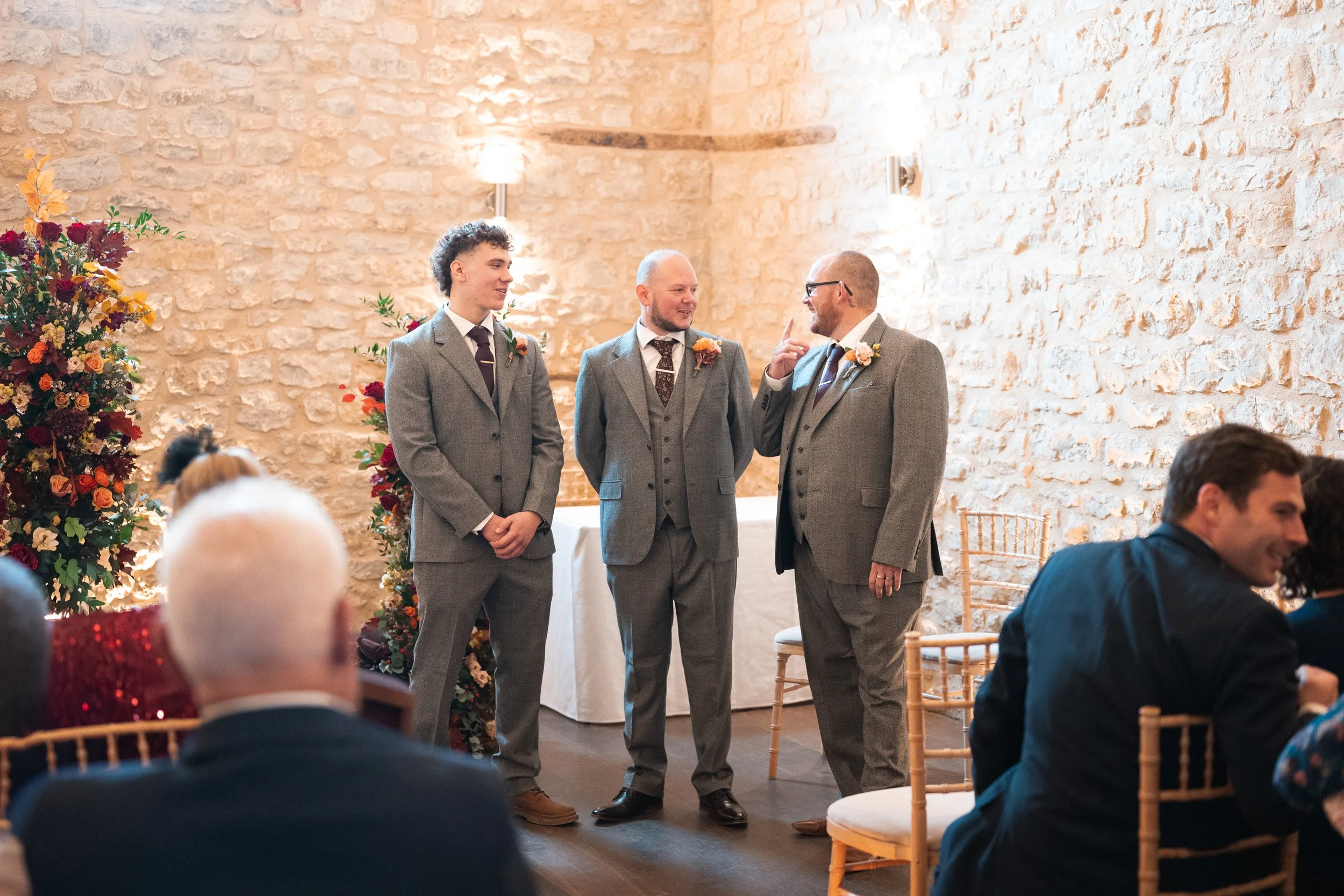 Three men in suits standing in front of a wedding ceremony, talking to each other, with guests seated around them in a room with stone walls.