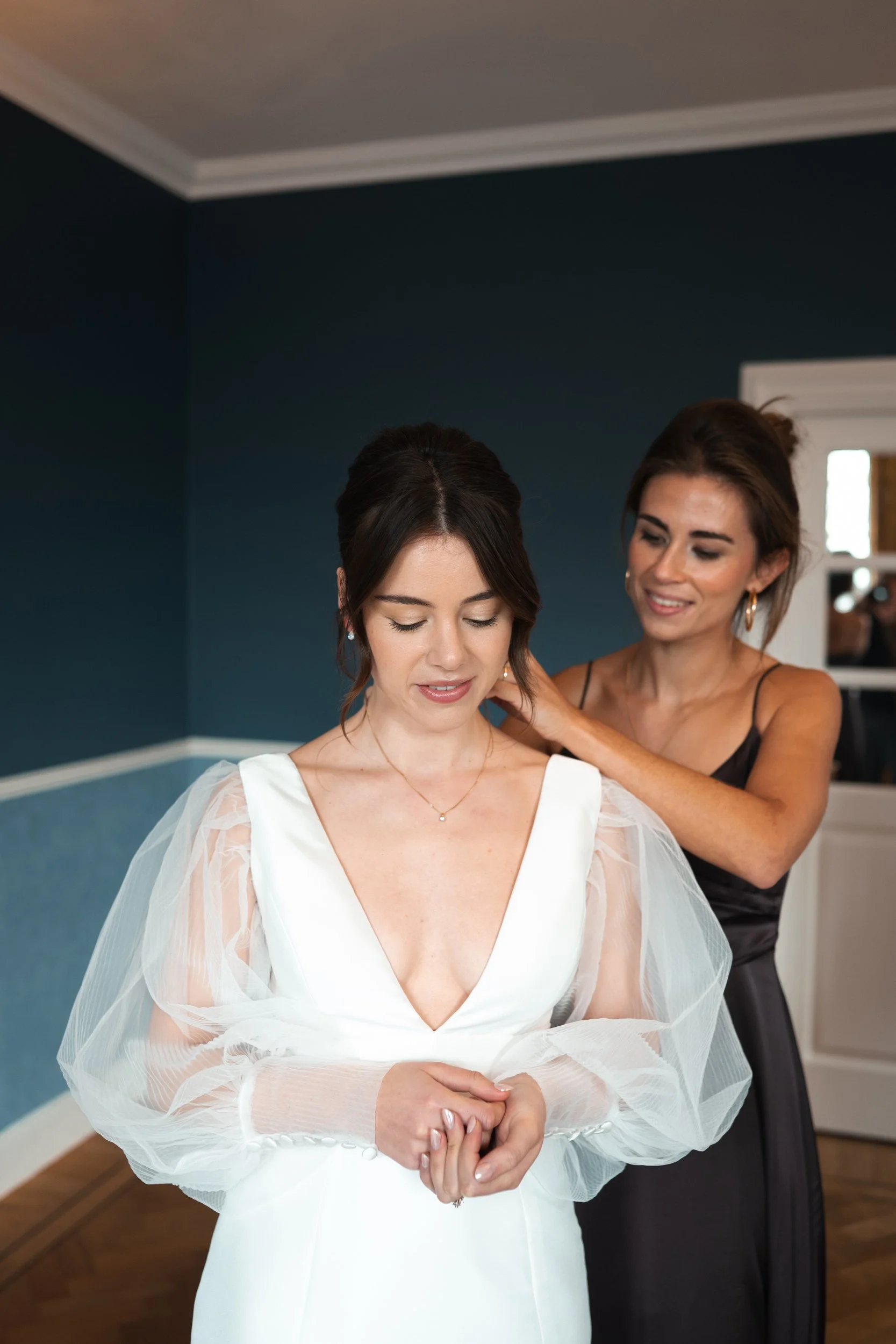 A woman dressed in a wedding gown with sheer, puffed sleeves during her wedding preparations, assisted by another woman in a black strap dress.
