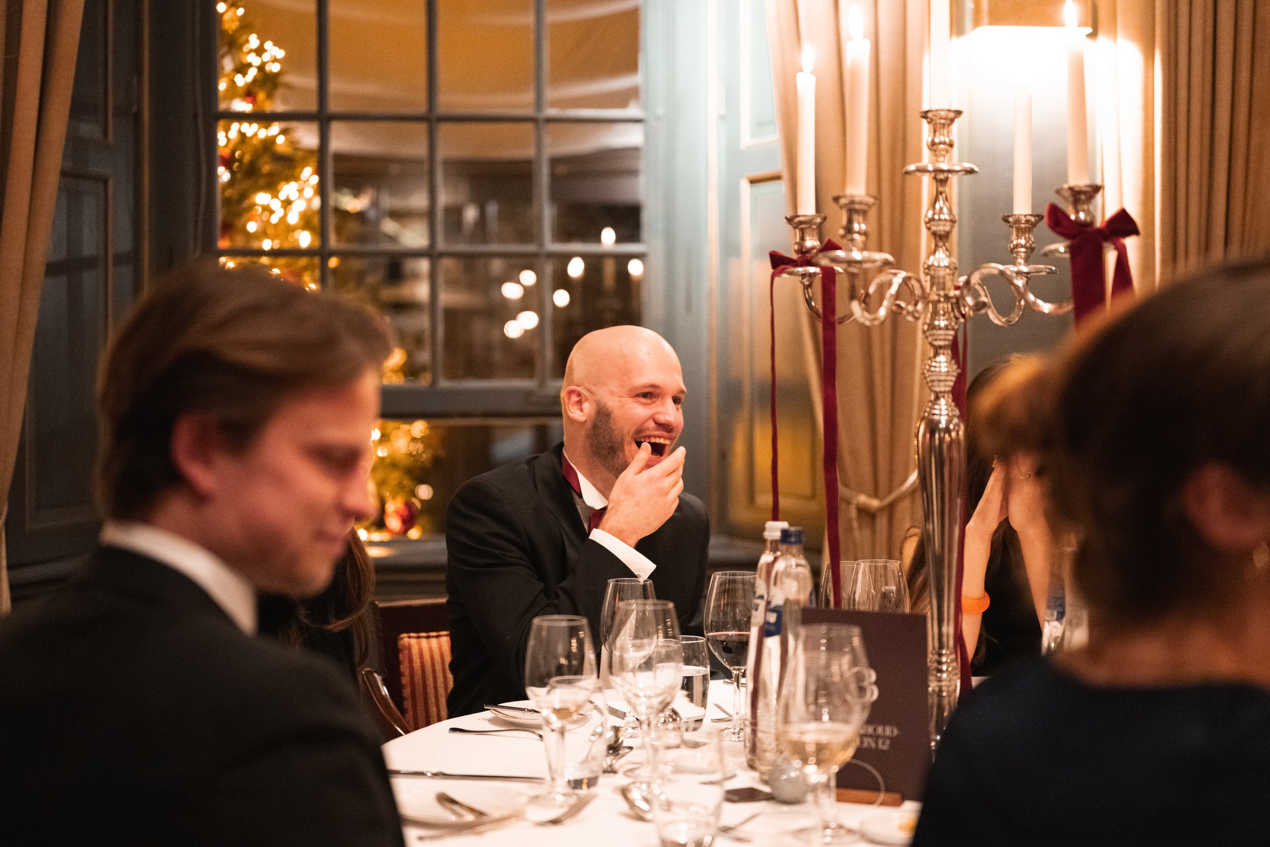 A man in a tuxedo sitting at a decorated evening dinner table, laughing and covering his mouth with his hand, with other guests blurred in the foreground. The setting is festive, with a candelabra and a Christmas tree in the background.