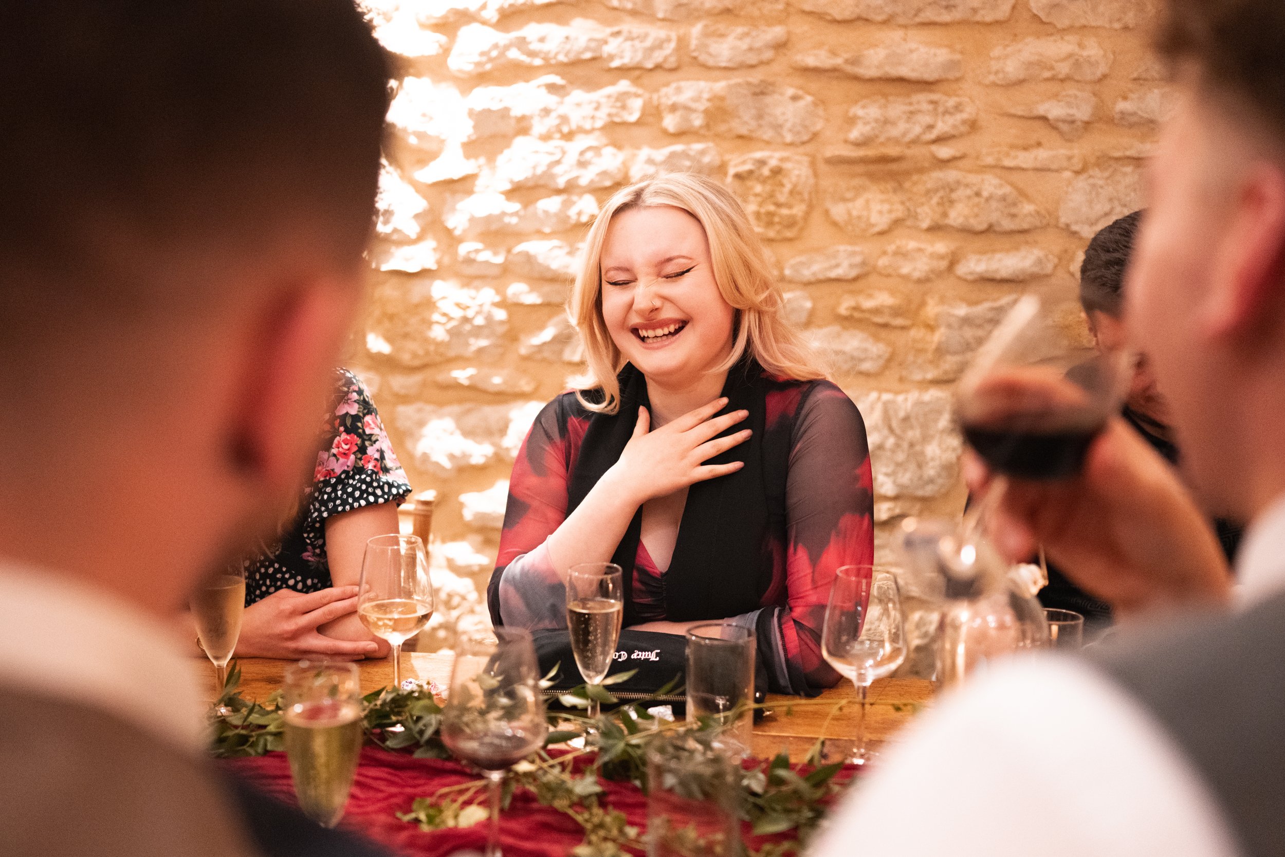 A group of friends at a dinner table, laughing and smiling, with glasses of wine and a decorated table in a cozy, rustic setting with a brick wall background.