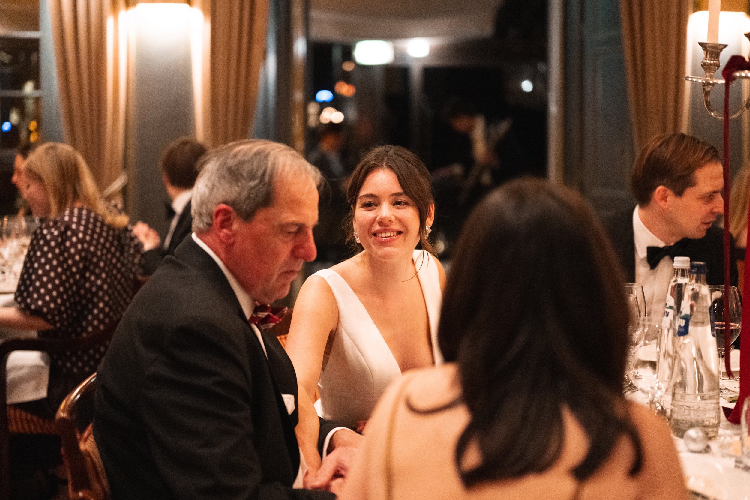 People dressed formally at a dinner party in an elegant restaurant, with a woman smiling in the center.