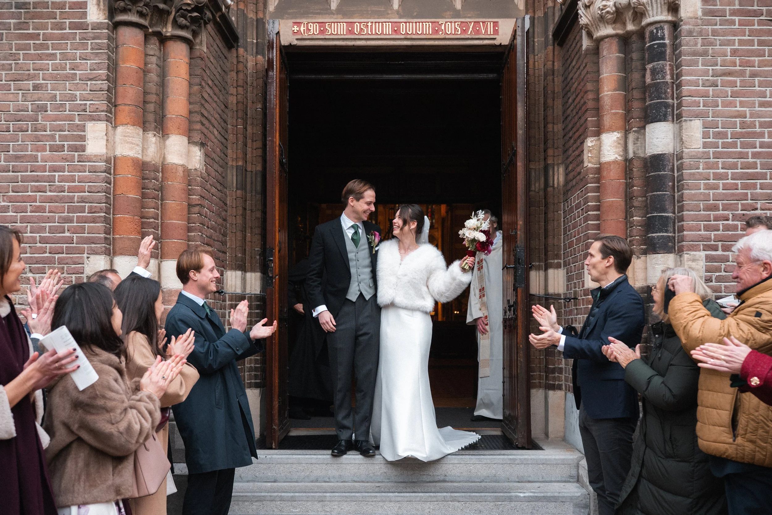 A newly married couple standing at the entrance of a church, smiling at each other, with guests clapping around them.