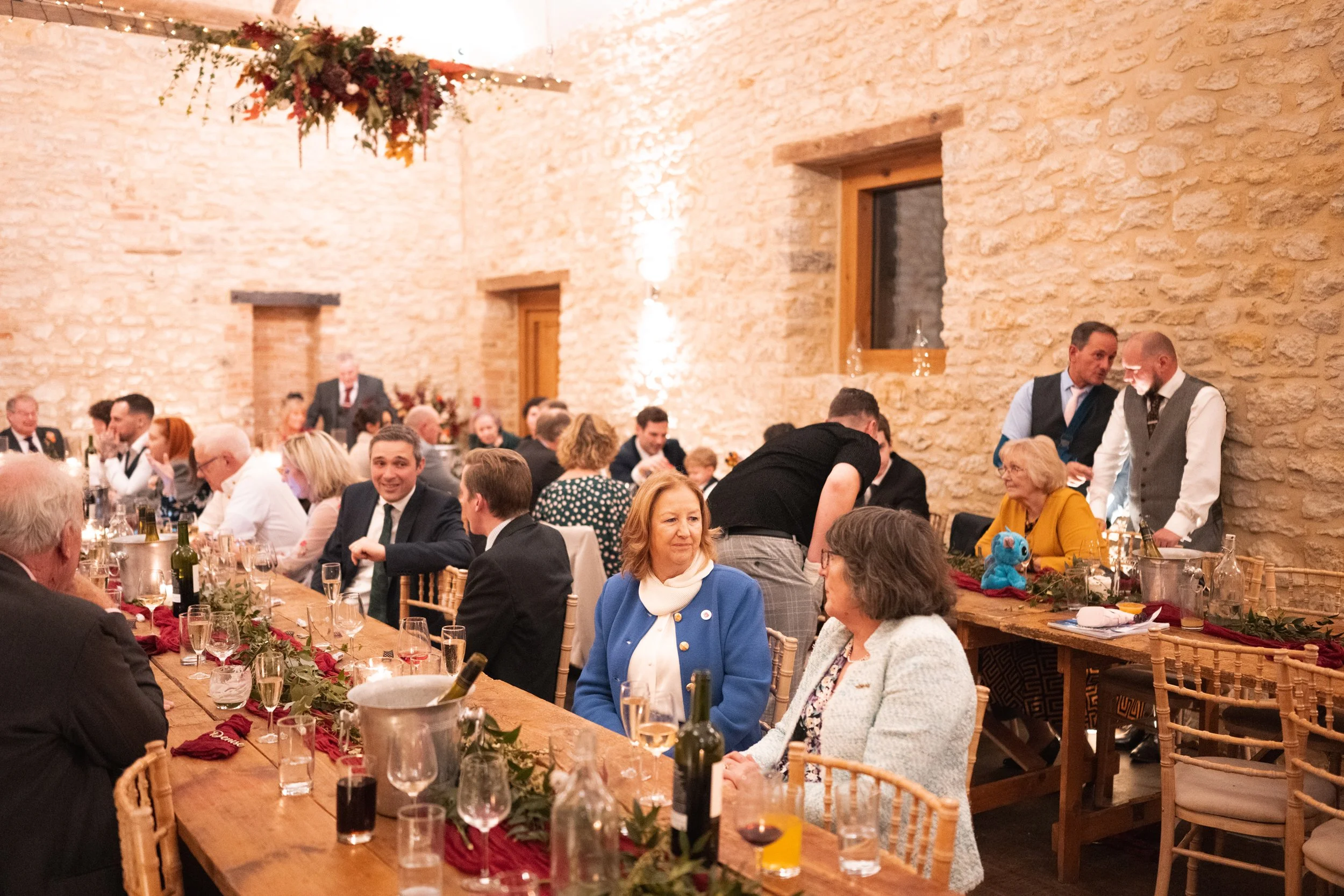 People gathered at a festive dinner party in a rustic stone-walled venue, with long tables decorated with greenery and red accents, and attendees engaged in conversation.